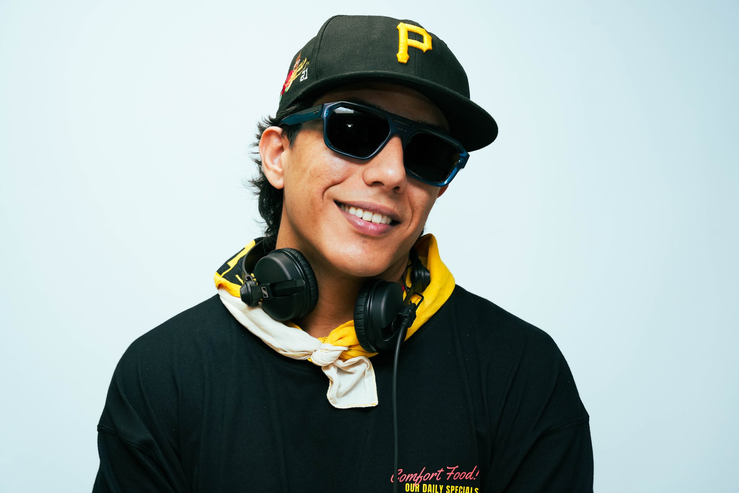 A smiling man wearing sunglasses, a black cap with a yellow 'P', a black T-shirt, a white and yellow neck scarf, and headphones around his neck.