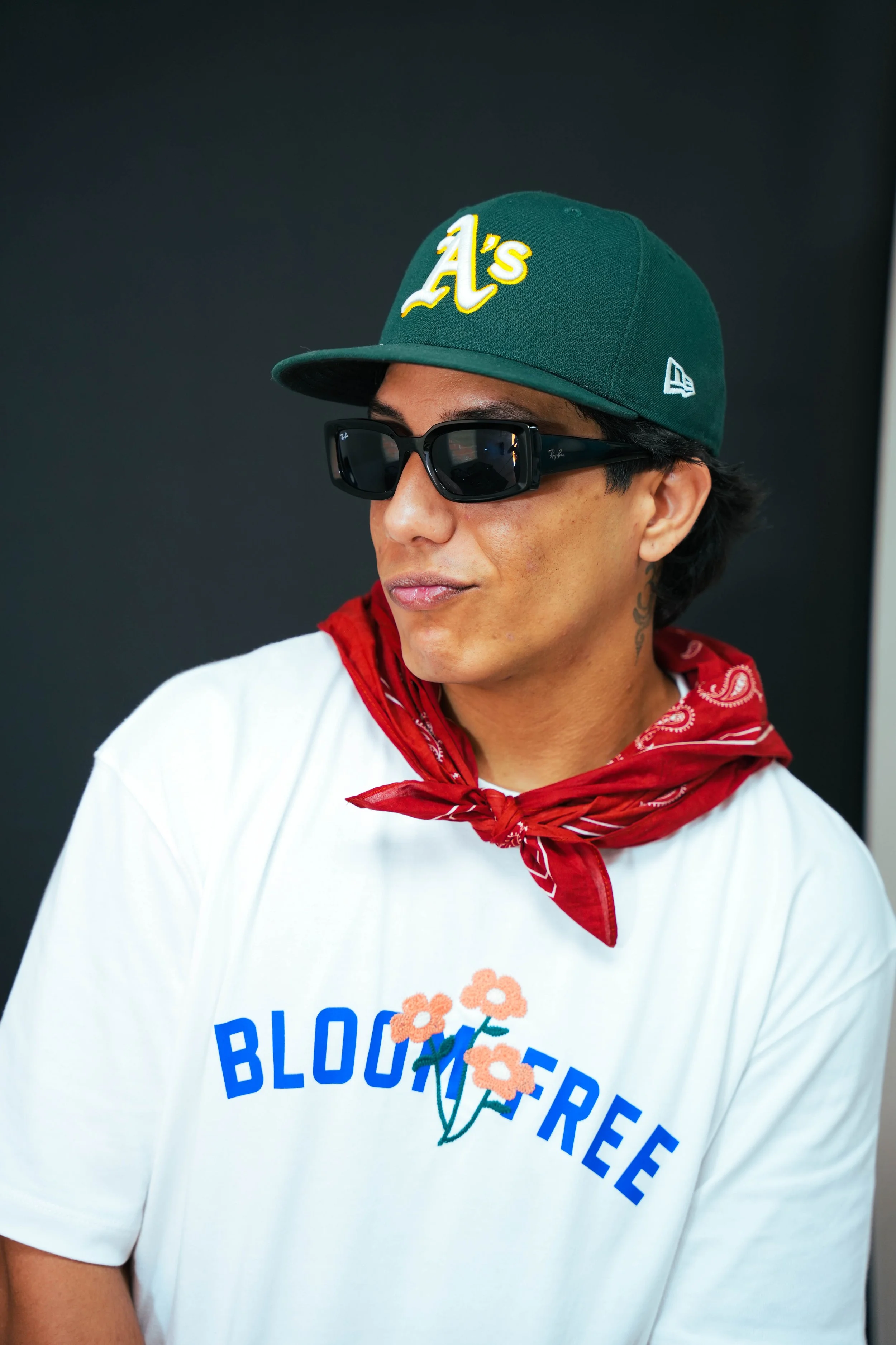 A young man wearing a green Oakland Athletics baseball cap, black sunglasses, a red bandana around his neck, and a white T-shirt with a flower graphic and the words 'BLOOMFREE' printed on it.