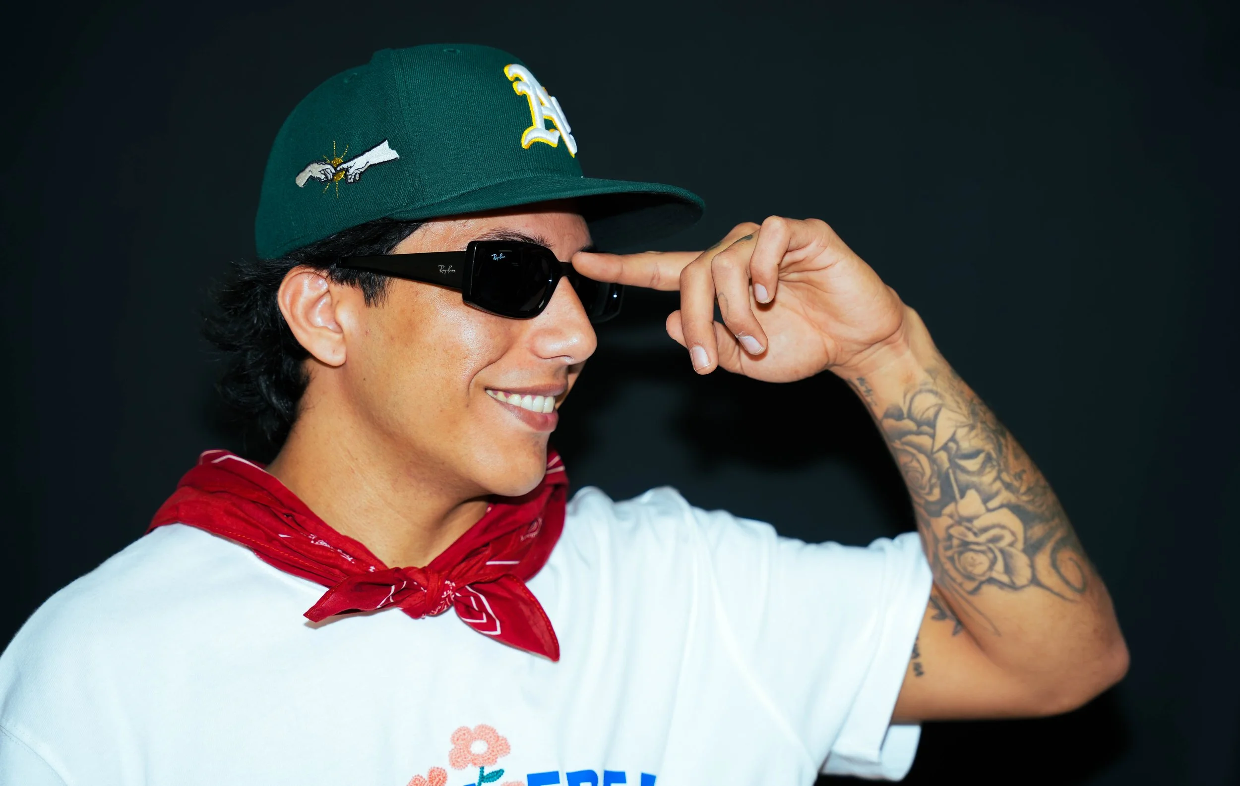 A person wearing a green baseball cap with embroidered patches, black sunglasses, a white T-shirt with logos, and a red bandana around their neck. They are smiling and saluting with their right hand, which has tattoos on the arm.
