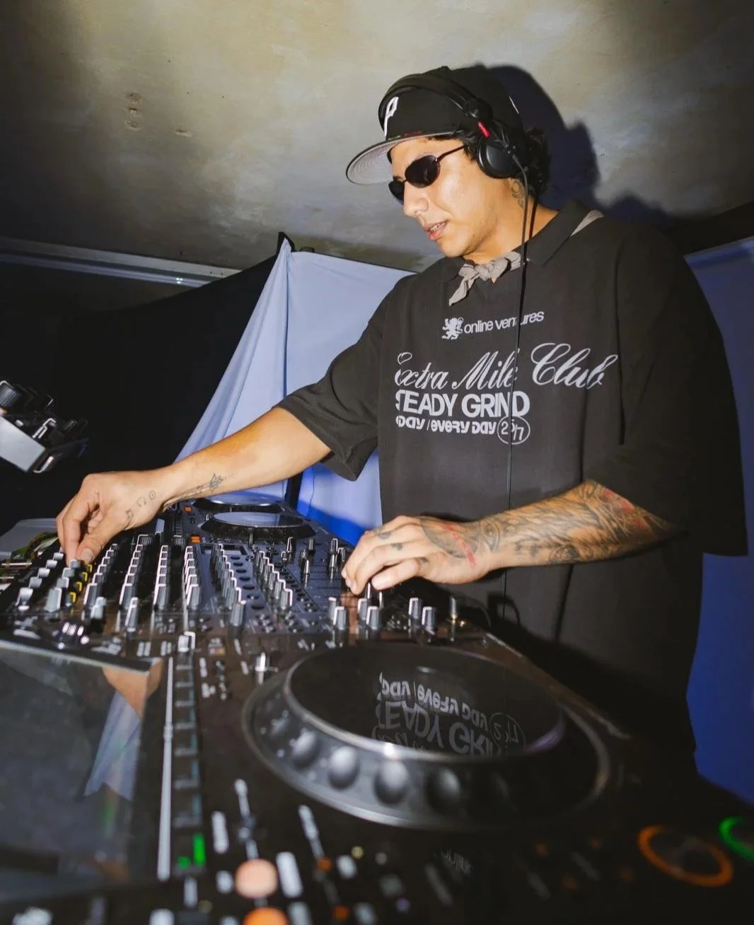 DJ wearing black sunglasses, black cap, and black T-shirt mixing music on DJ equipment in a dimly lit setting.