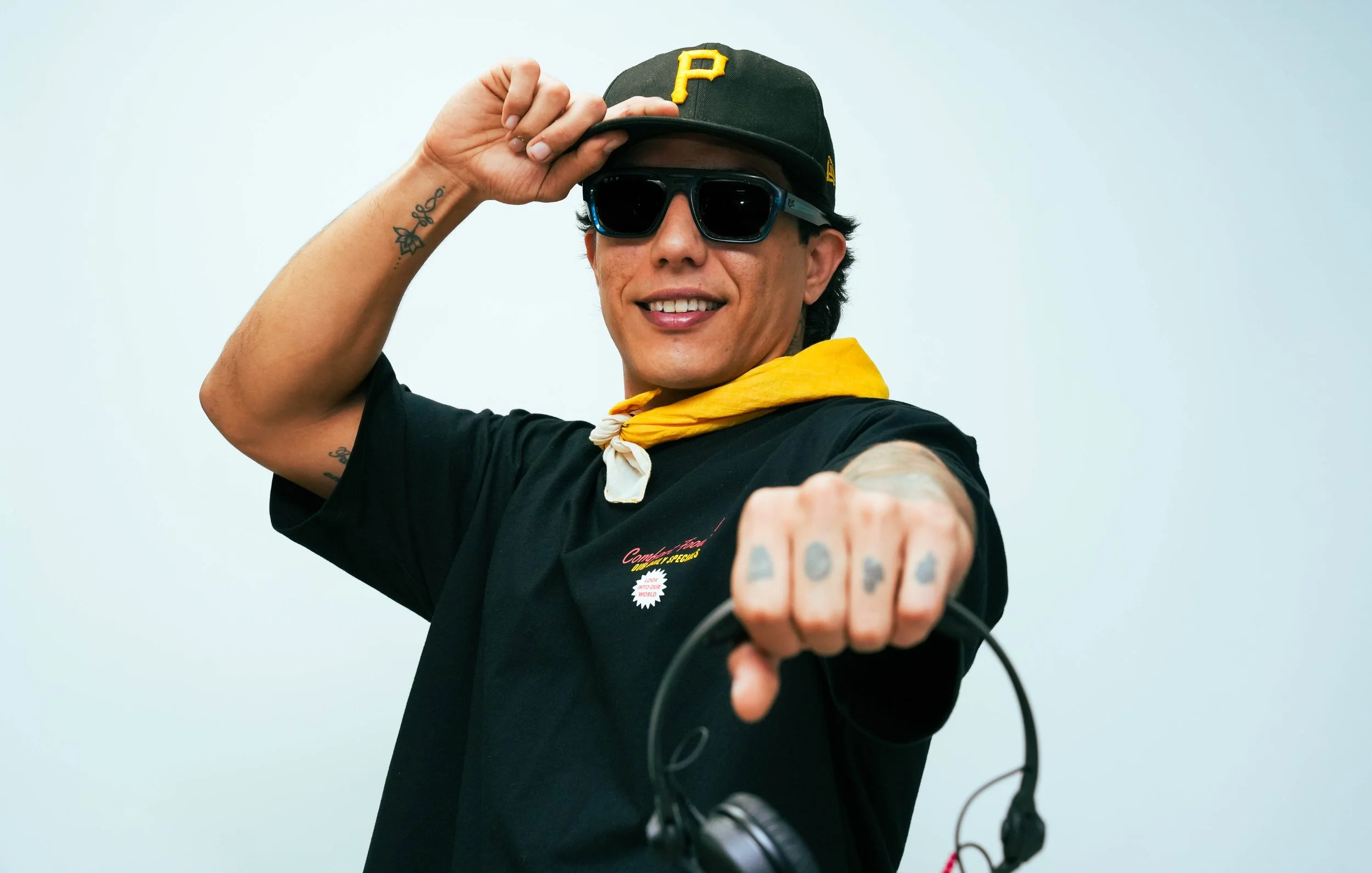 A man wearing sunglasses, a black baseball cap with a yellow 'P', a black T-shirt, and a yellow bandana around his neck is smiling and pointing towards the camera. He has tattoos on his arm and hand, and is holding headphones.