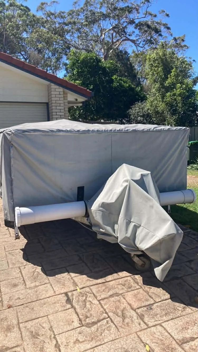 A large pop-top caravan with a Hamilot cover, situated on a paved surface with trees and a house with a red roof in the background.