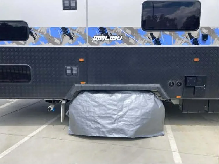 A Malibu motorhome parked in a parking lot with a gray Hamilot cover over the tire, and the upper motorhome painted in a blue, black, and white design.