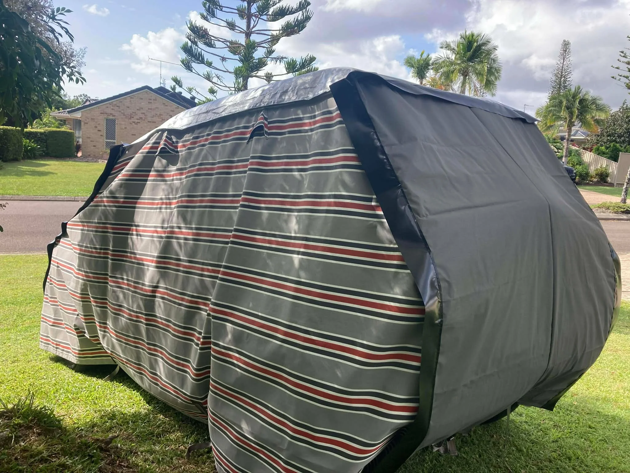 A striped outdoor Hamilot Cover with a red and grey striped Hamilot cover on a Suzuki Jiminy set up on a lawn with a residential neighborhood, trees, and a cloudy sky in the background.