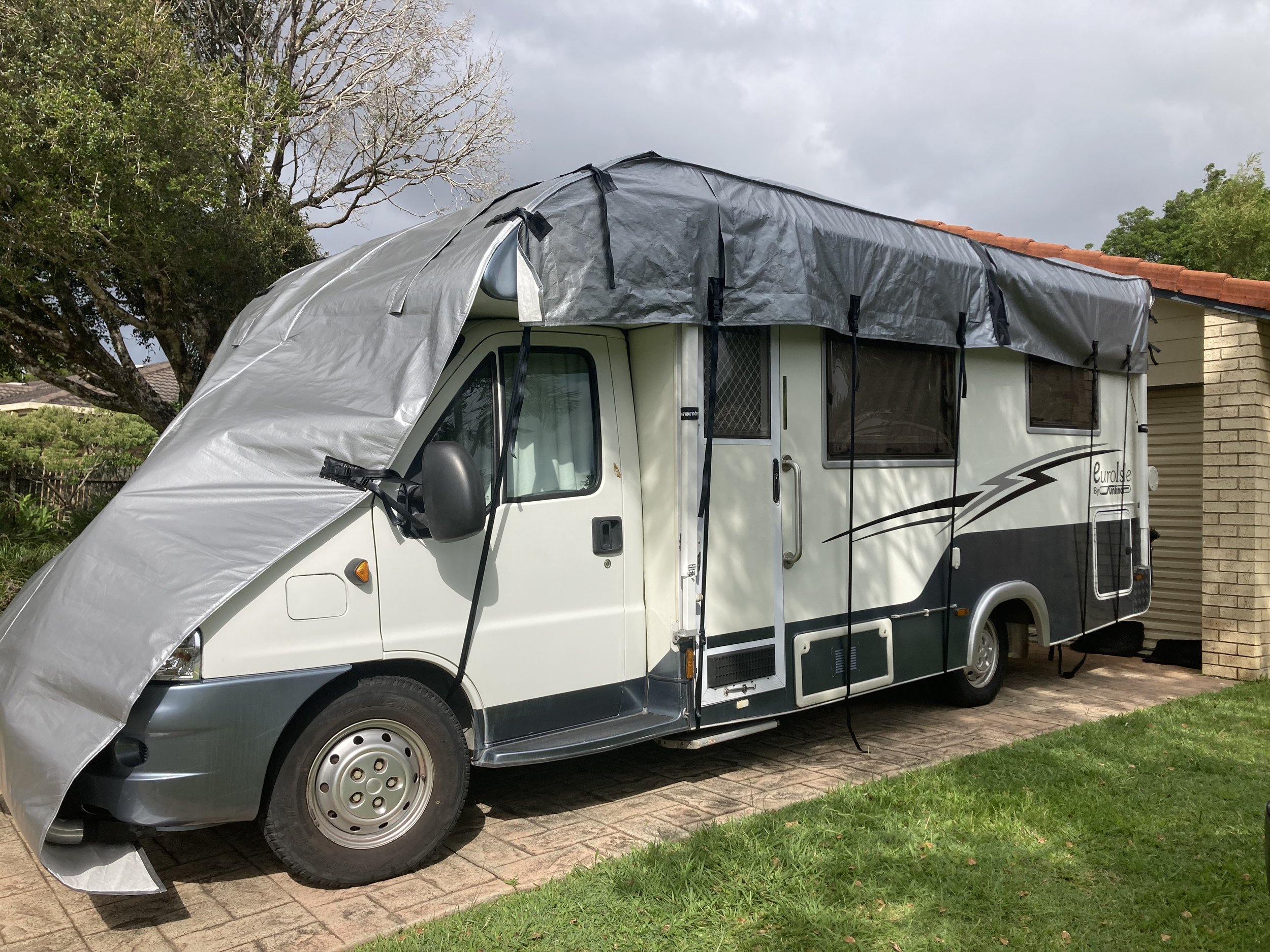A camper van with an attached gray tarp, parked on a brick driveway next to a brick house with a red tile roof, green grass, trees, and a cloudy sky in the background.