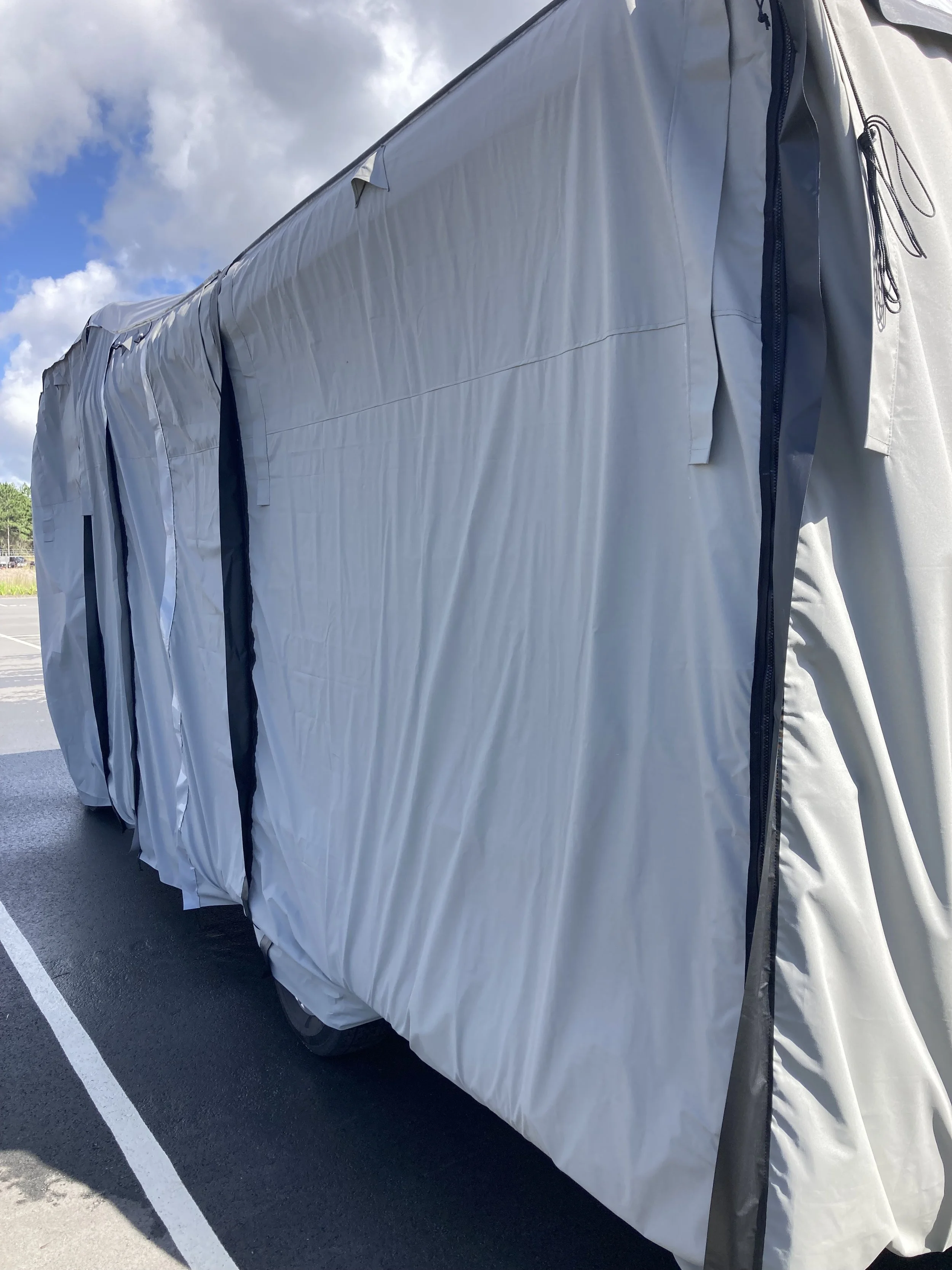 Large vehicle covered with a gray protective tarp, parked in a parking lot under a partly cloudy sky.