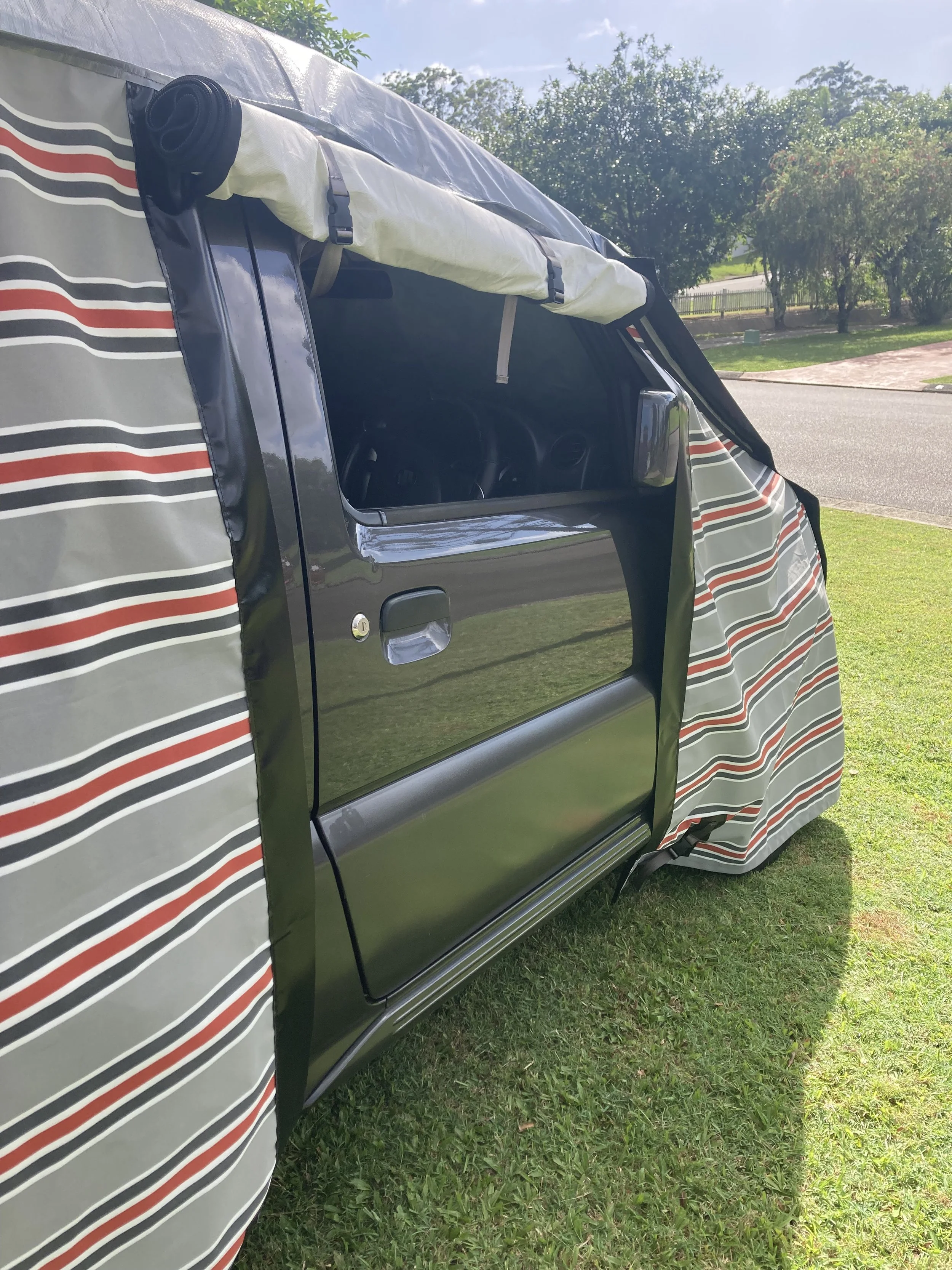 Side view of a two-door Jimny with a striped Hamilot hail cover attached to it, parked on grass near a street with trees in the background.