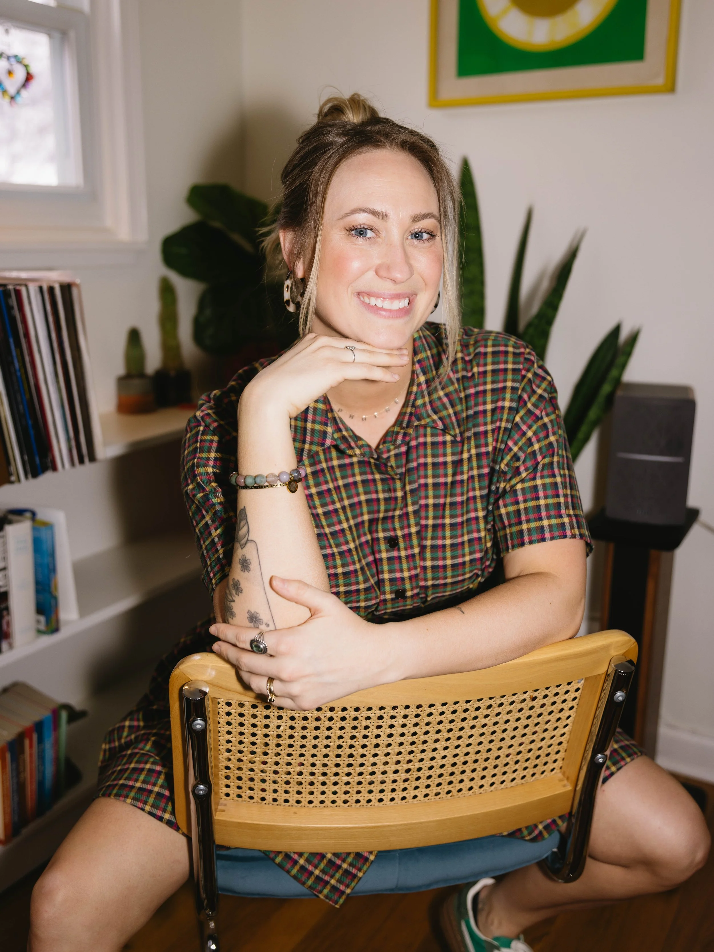 A woman sitting on a wooden chair with a woven backrest, smiling and posing with her hand on her chin, in a room with books, plants, and wall art.