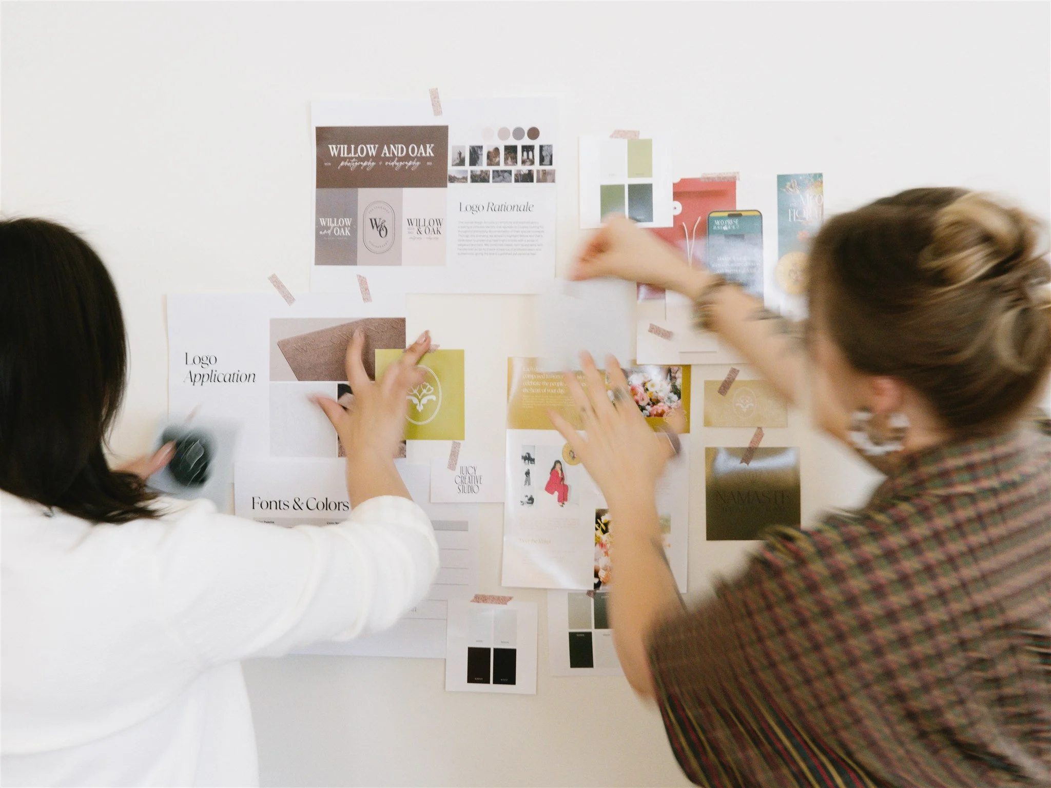 Two women working on a project, taping and arranging images and notes on a white wall for a design or branding presentation.