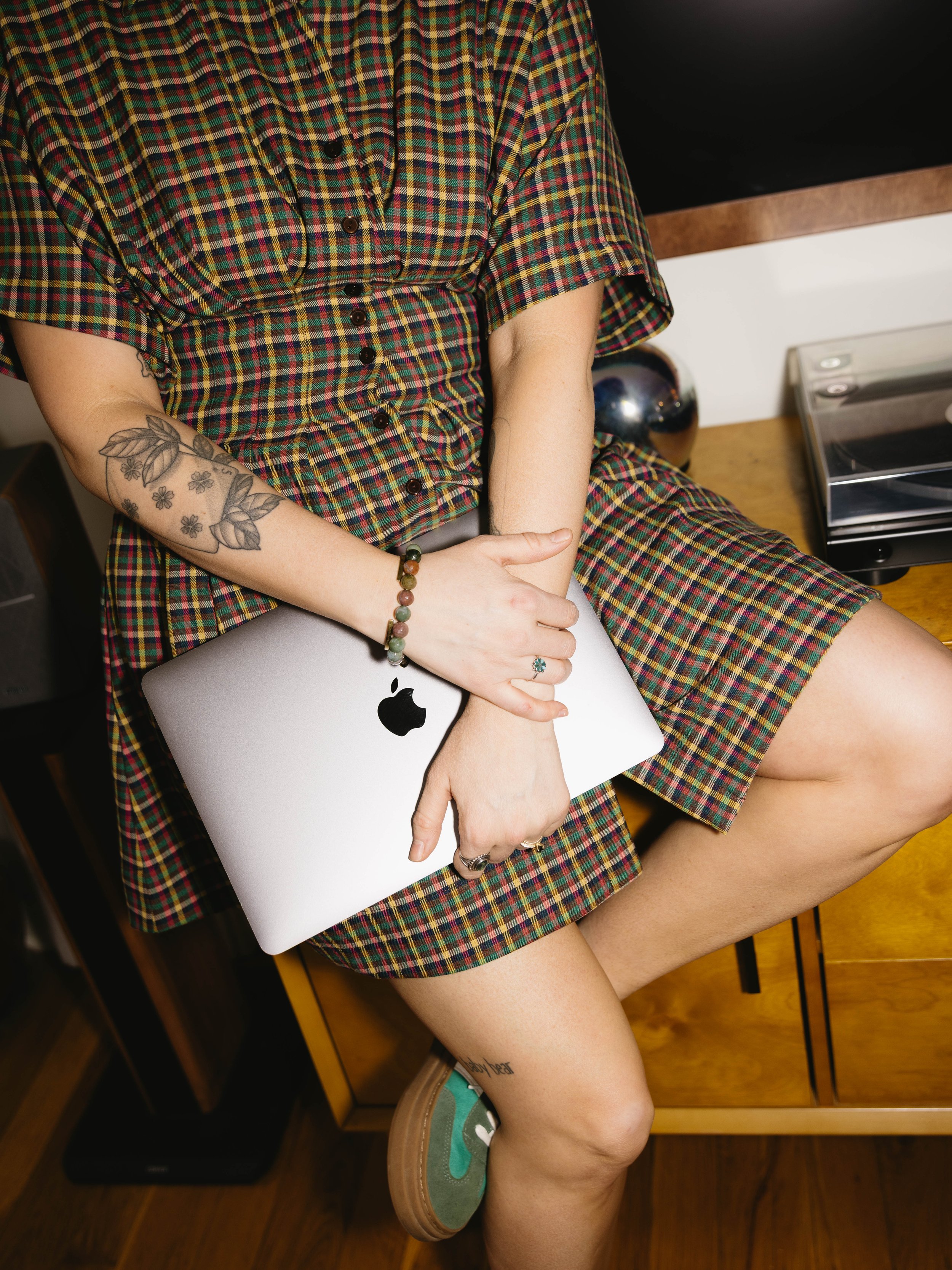 Person wearing a plaid dress sitting on a wooden desk, holding a closed silver MacBook laptop, with tattoos on their arm and leg, wearing rings and a beaded bracelet.