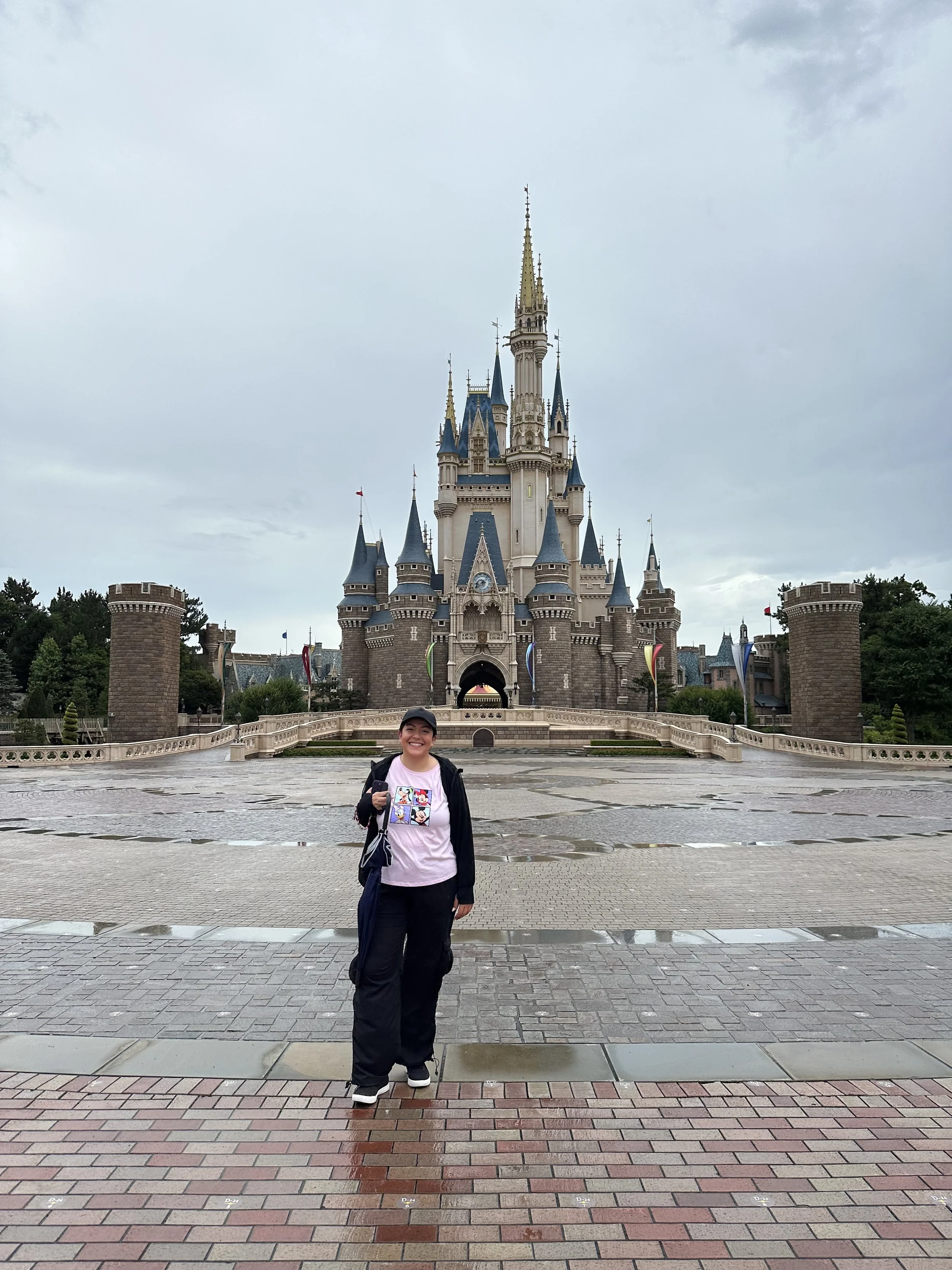 A woman standing in front of a castle at Disneyland, smiling, wearing a pink shirt, black pants, and a black jacket, with overcast skies.