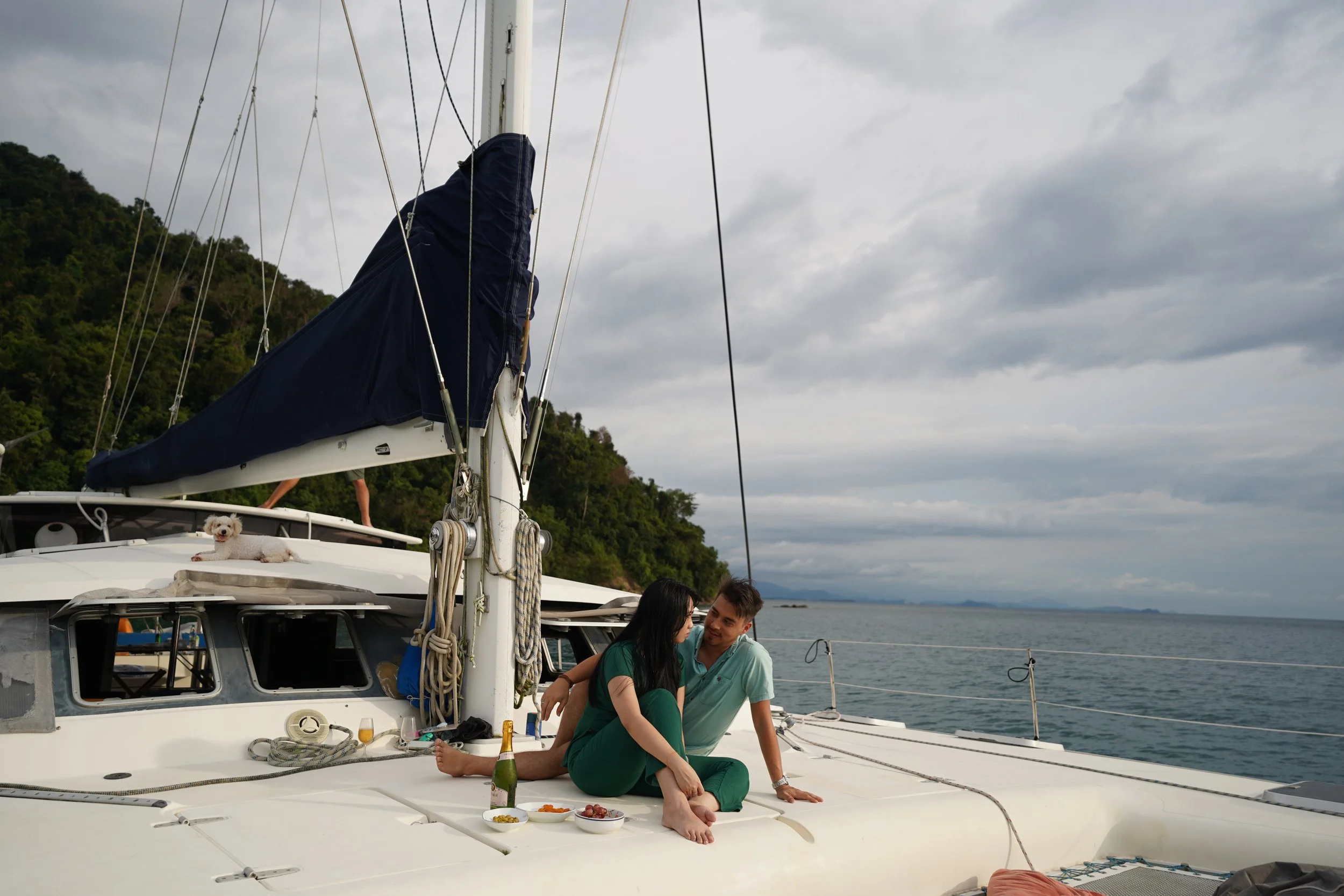 A man and woman sitting on the deck of a sailboat, smiling and looking at each other. The woman is in green clothing, and the man is in a light shirt. A small dog is lying on the roof of the boat, and there are snacks and drinks nearby.