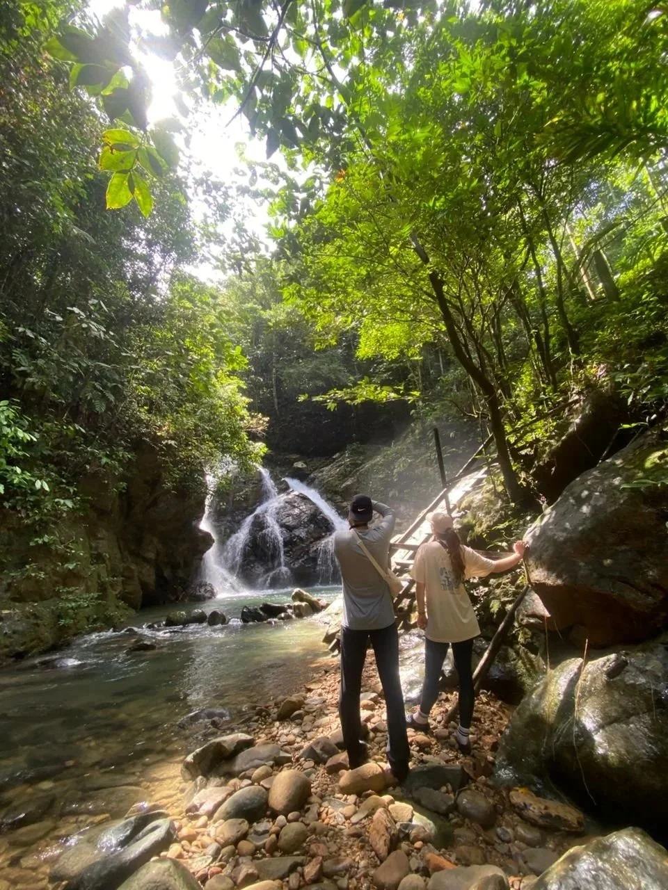 Two people hiking near a small waterfall in a lush, green forest, with sunlight filtering through the trees.