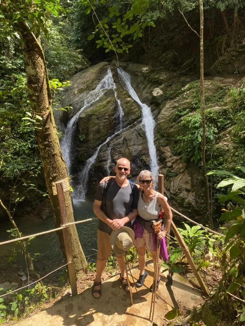 A man and woman stand on a dirt path in front of a waterfall in a lush green forest. They are smiling, with the man holding a hat and the woman holding a walking stick. The setting appears to be a nature trail with a wooden fence and rope barrier.