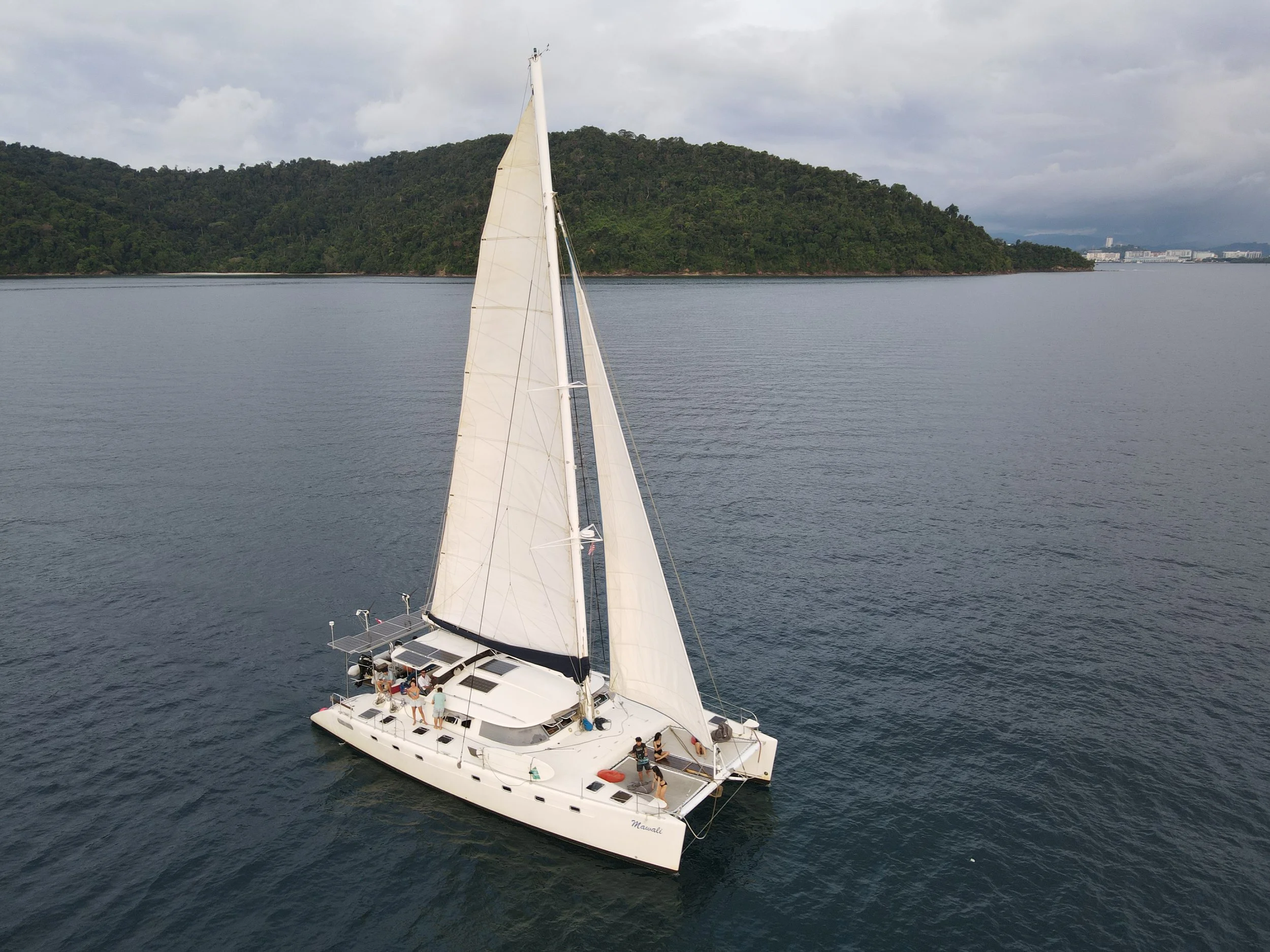 A white sailing catamaran with multiple sails on calm water near a green, hilly island with a cloudy sky.