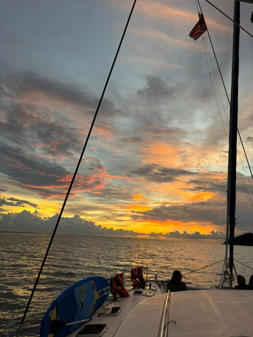 Sunset over the ocean viewed from a sailboat deck, with a small American flag on a pole and people sitting at the bow.