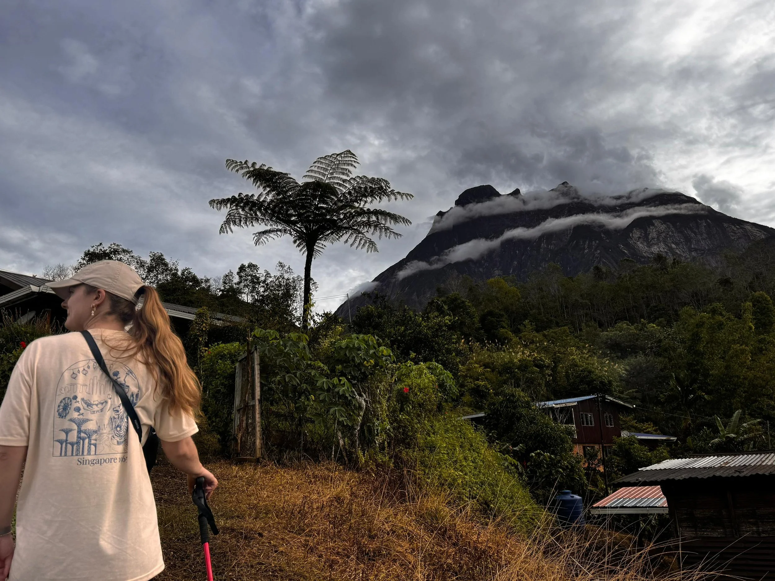 A woman with red hair, wearing a beige cap and T-shirt, holding hiking poles, looking away. She is standing outdoors in a mountainous area with dense trees, a hillside with small houses, and a dark cloudy sky overhead.