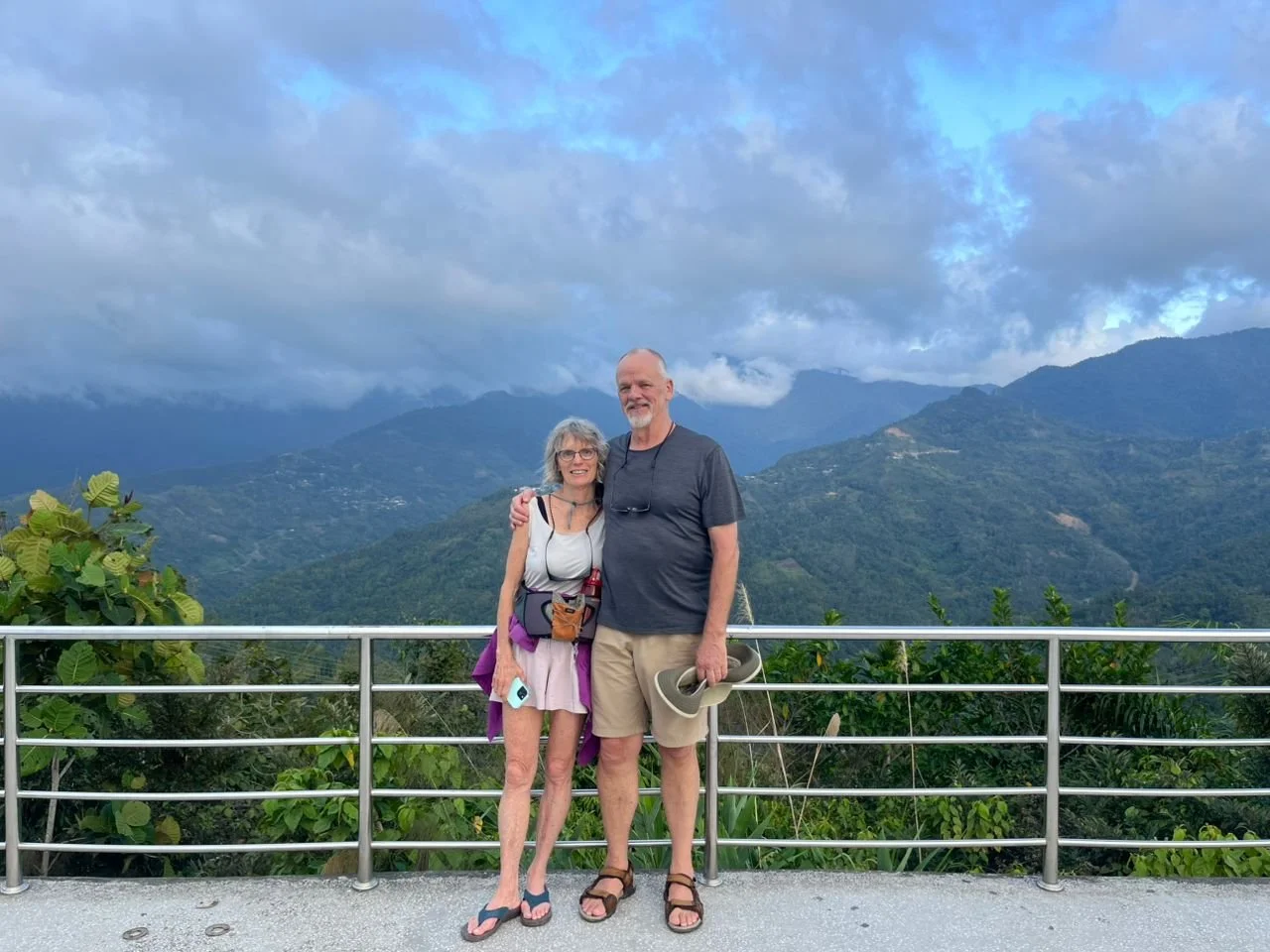A smiling elderly couple standing on a viewing platform with a mountain landscape in the background, the woman wearing a light dress and glasses, and the man wearing a dark T-shirt and shorts, holding a hat.