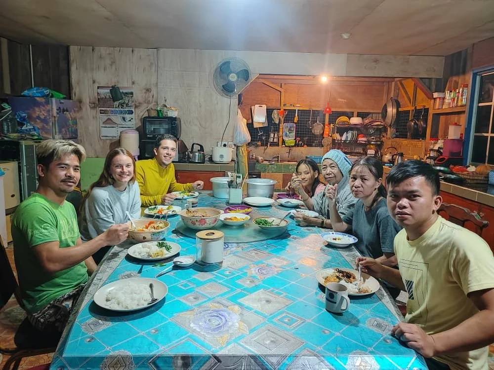 A group of eight people sitting around a dining table with plates of food, in a rustic kitchen.