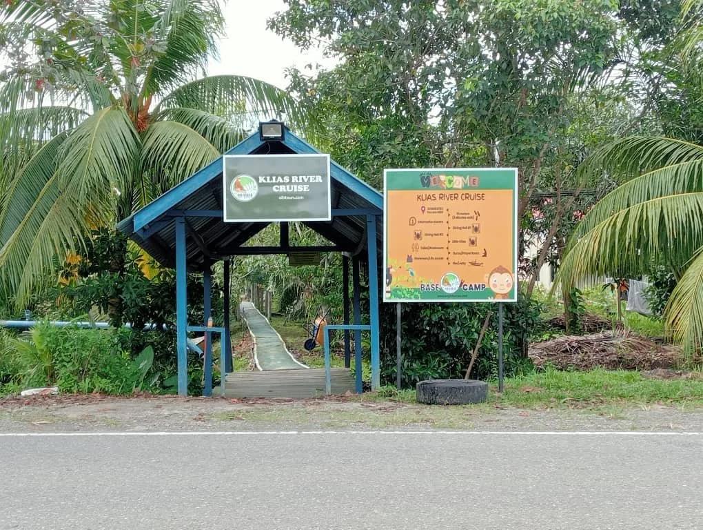 Entrance to Kailas River Cruise with a blue small gateway and a sign. There are lush green trees and plants around, with a narrow pathway leading into the background.