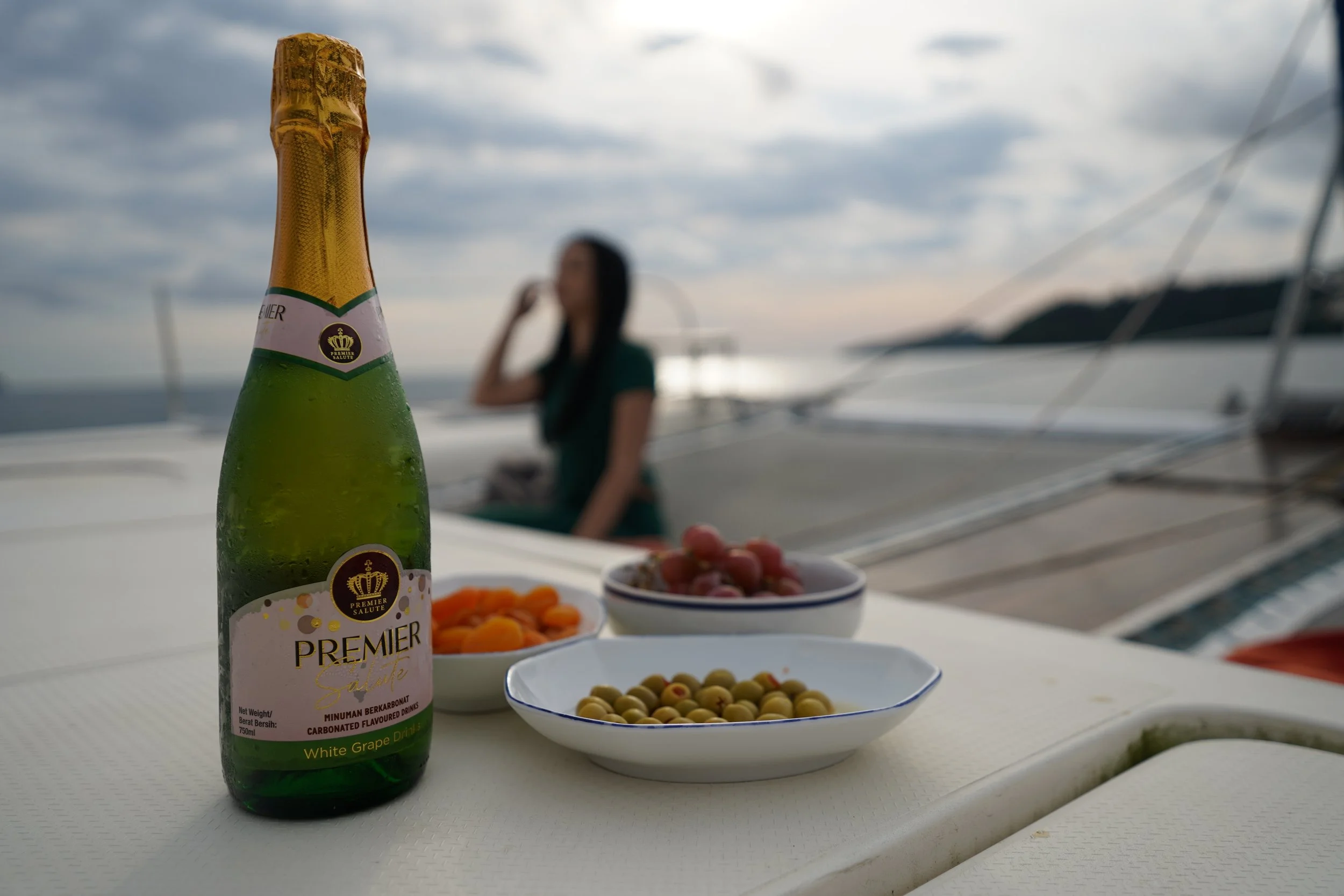 A bottle of Premier Sparkling White Grape Drink with bowls of snacks on a table on a boat. A woman is sitting in the background with a blurred view of the water and sky.