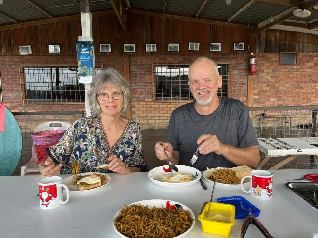 A smiling elderly man and woman sitting at a table outdoors, eating Asian-style dishes including noodles and eggs with toast, in a rustic setting with brick and wood background.