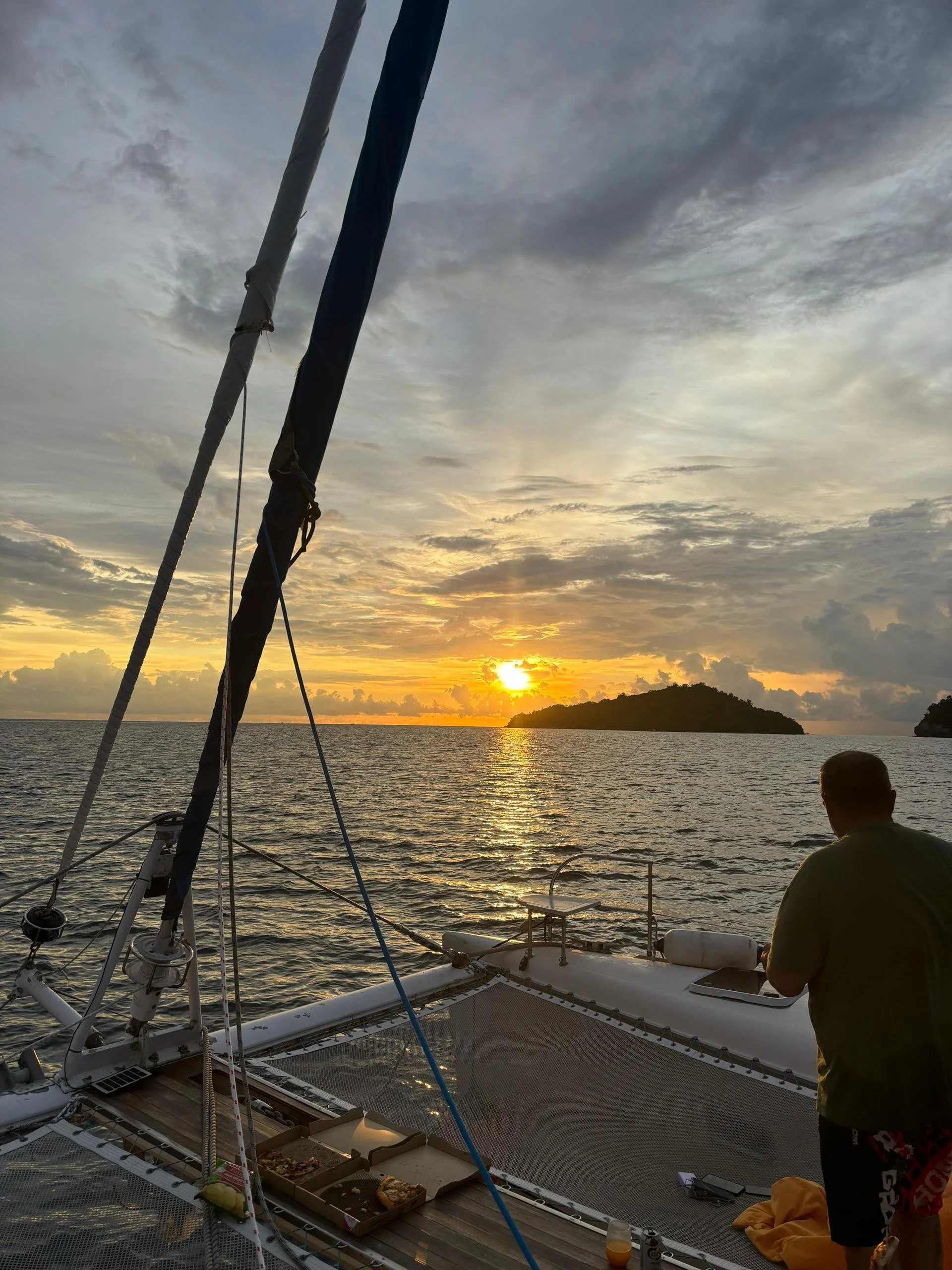 A person on a sailboat looking at a sunset over the ocean with islands in the distance, a pizza box and drinks on the boat.