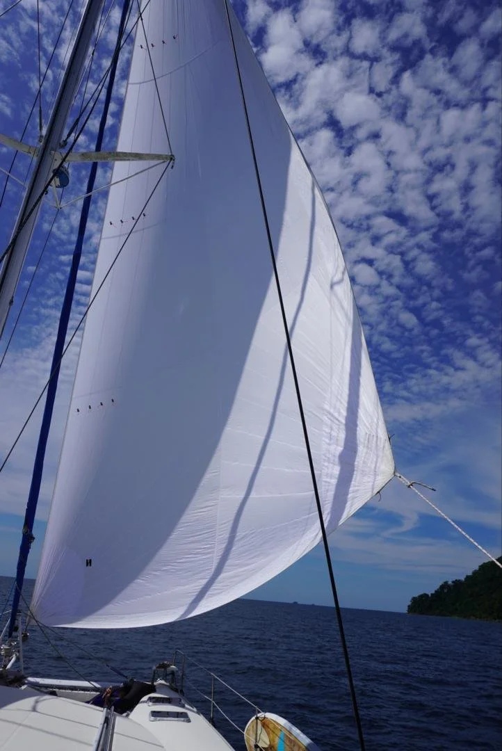 A sailboat with a large white sail on the open ocean, with a partly cloudy sky and a distant landmass.