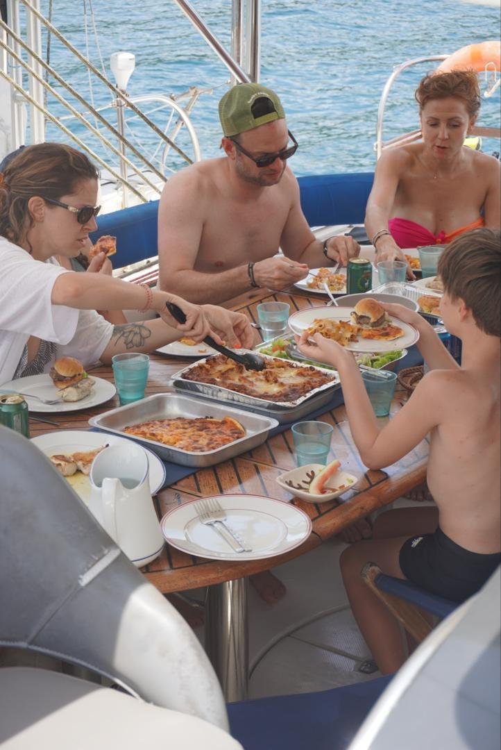 Group of five people having a meal on a boat, eating pizza and other food, with water in the background.