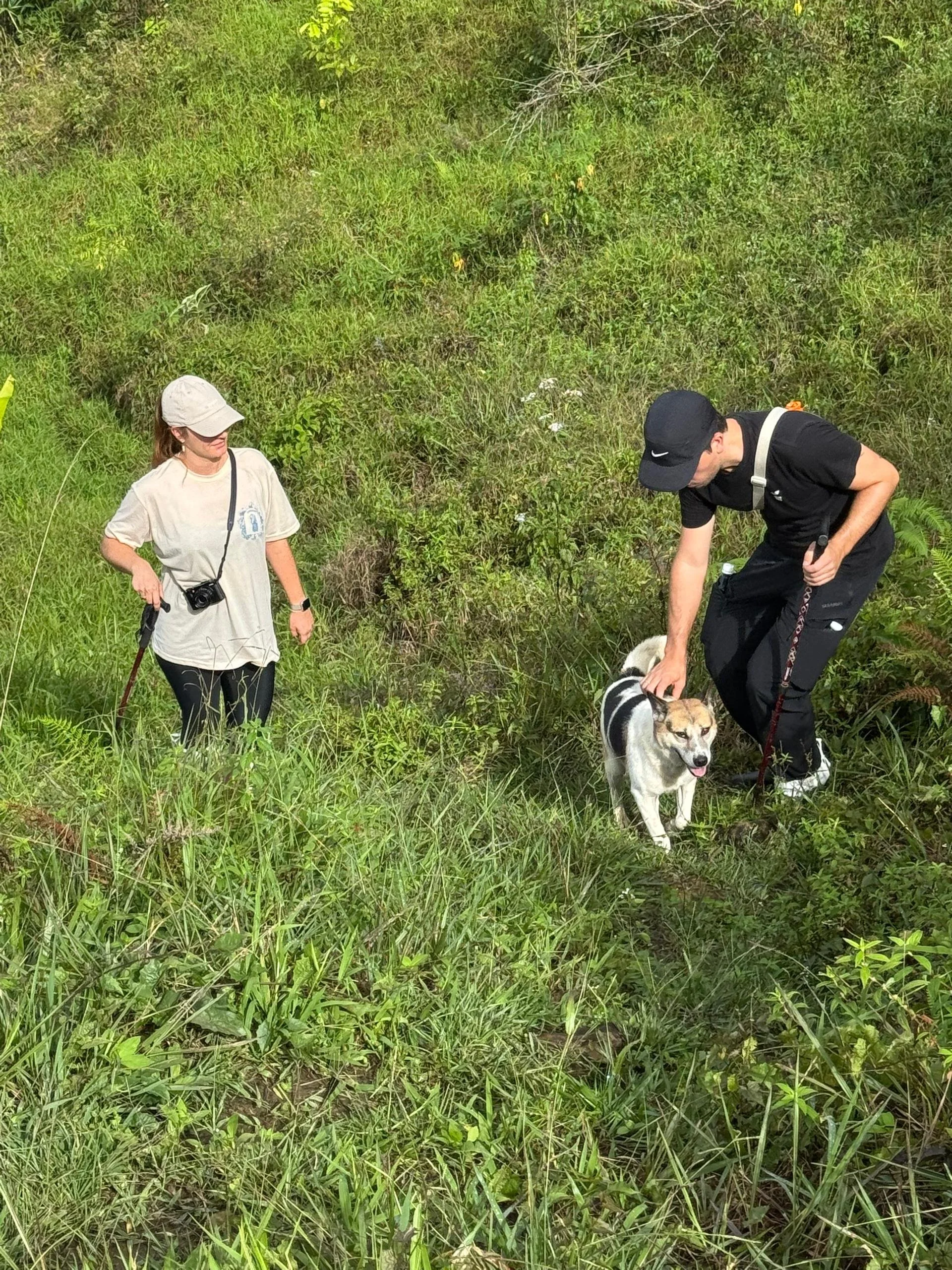 Two people hiking through grassy terrain with a dog. One woman wears a beige hat and large camera, the man wears a black cap and monofilament. The dog is a small to medium breed with a tan and white coat, standing on a narrow trail.