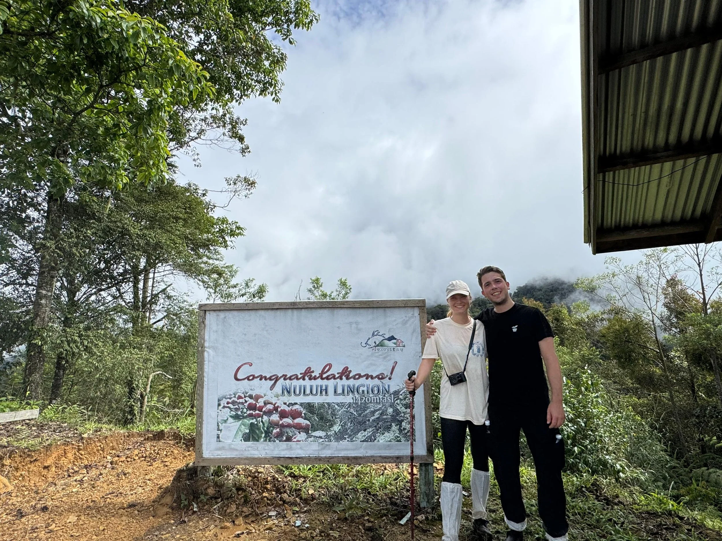 Two smiling hikers, a woman and a man, stand together outdoors in front of a sign that says "Congratulations! Nuluh Lincoln 1420 m asl". They are surrounded by trees and mountainous landscape, with cloudy sky overhead.