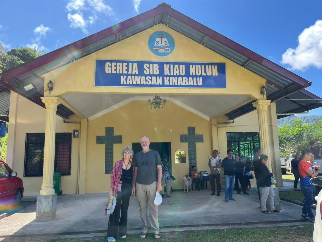 People standing outside a church with a large blue sign that reads "GEREJA SIB KIAU NULUH KAWASAN KINABALU" under a blue sky with a few clouds