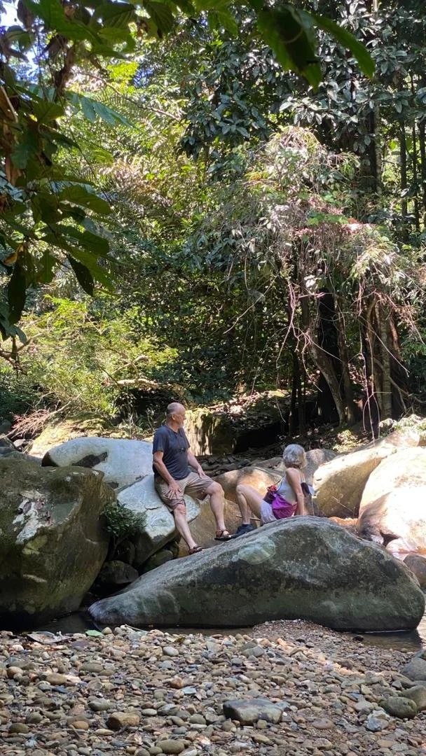 Two elderly people sitting on large rocks in a creek surrounded by dense trees and foliage at Murug Turug waterfall hike.