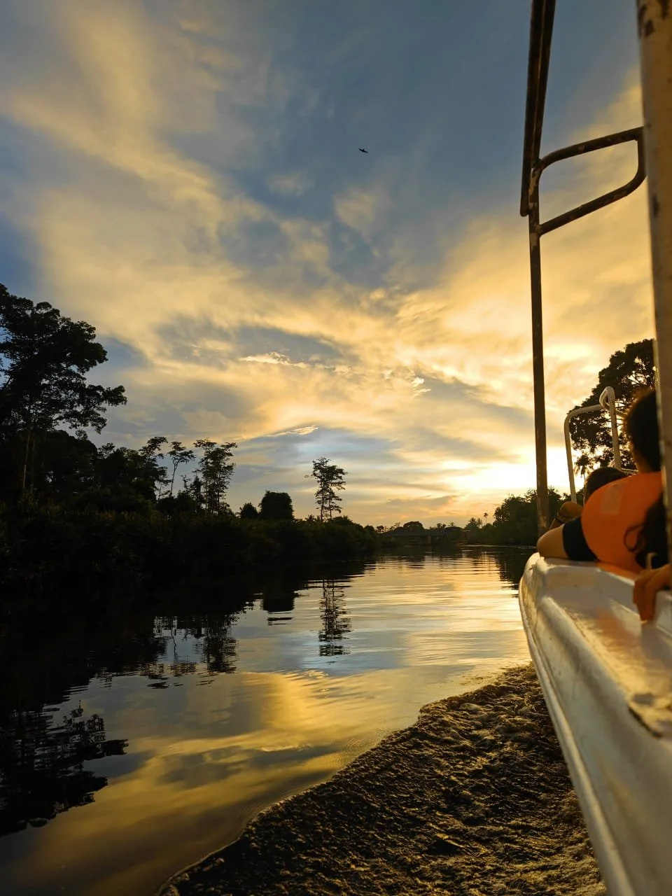 A river at sunset with trees on either side and a sky filled with clouds. Part of a boat with people on it is visible on the right side.