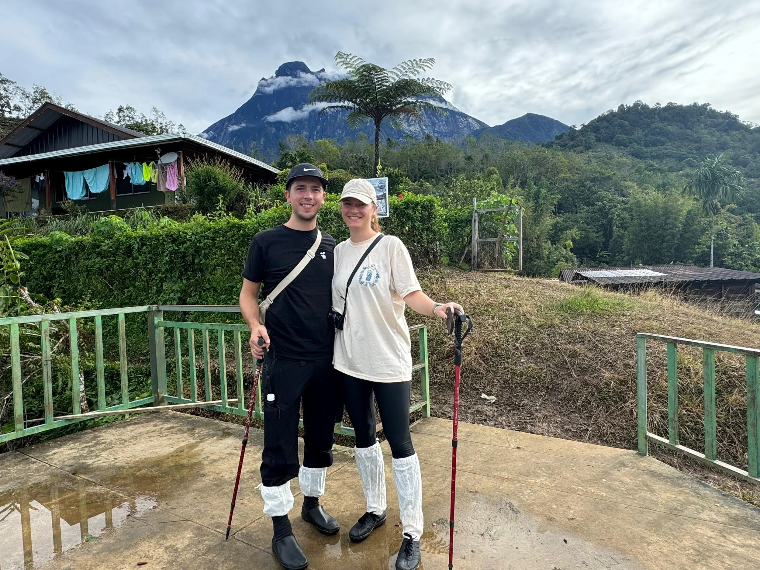 Two hikers, a man and a woman, standing on a concrete trail with hiking poles, smiling, surrounded by lush greenery and mountains in the background.