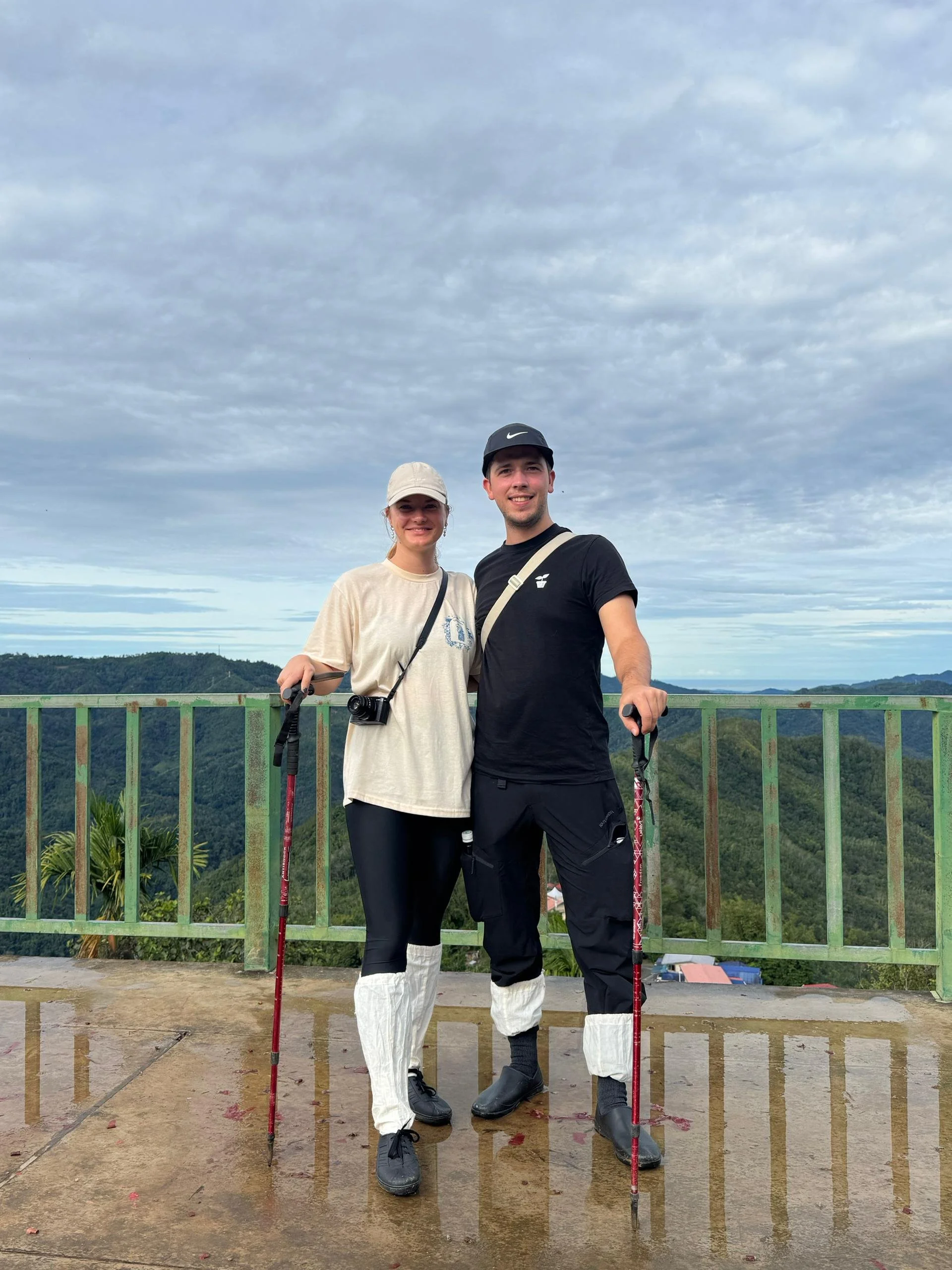 A smiling couple standing together on a viewing platform, holding trekking poles, with a scenic green mountain landscape in the background. They are dressed casually for hiking, with the woman wearing a beige hat and the man wearing a black cap.