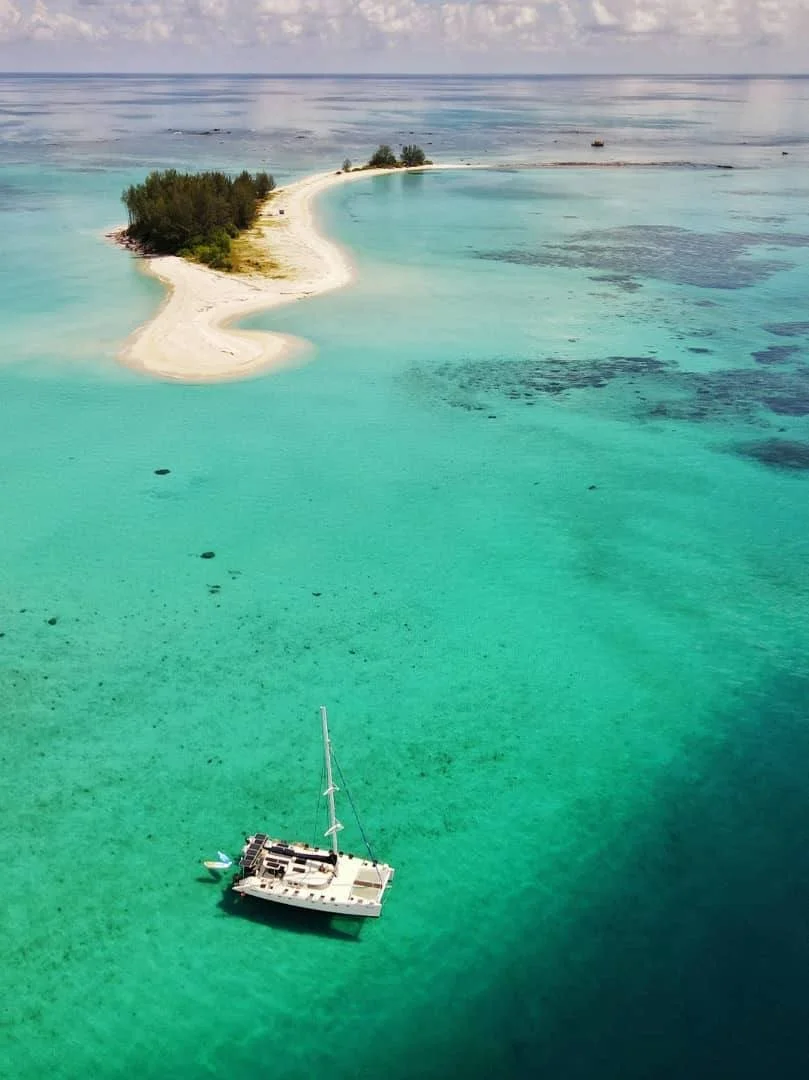 A sailboat floating in turquoise water near a sandy island with greenery, with a distant shoreline and cloudy sky in the background.