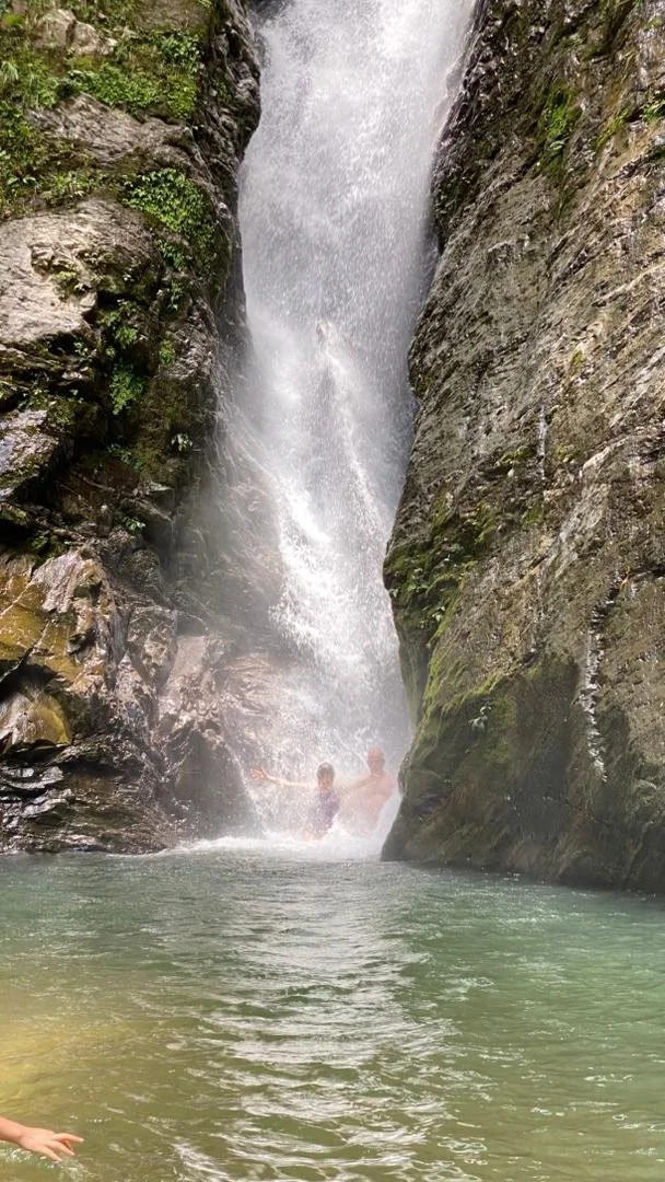 Two people swimming in a natural pool at the base of a waterfall between rocky cliffs with moss and small plants at Murug Turug Waterfall Hike.