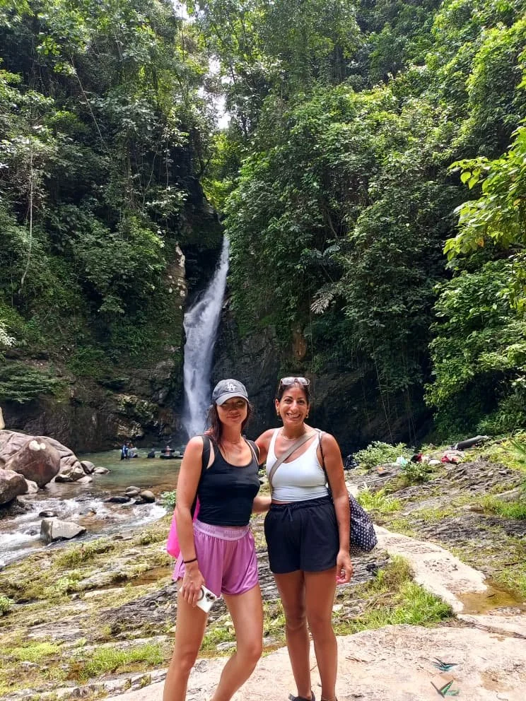 Two women standing in front of a waterfall in a lush green forest. One is wearing a black tank top, pink shorts, and a cap, and the other is wearing a white tank top and black shorts.