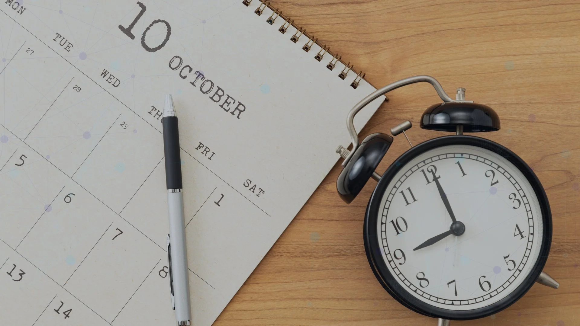 Alarm clock beside a calendar and pen on a wooden desk.