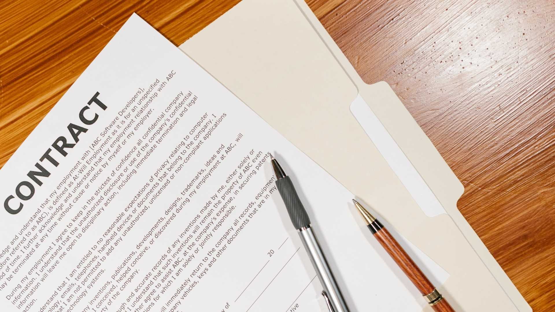 Contract document in an open folder with two pens on a wooden desk.