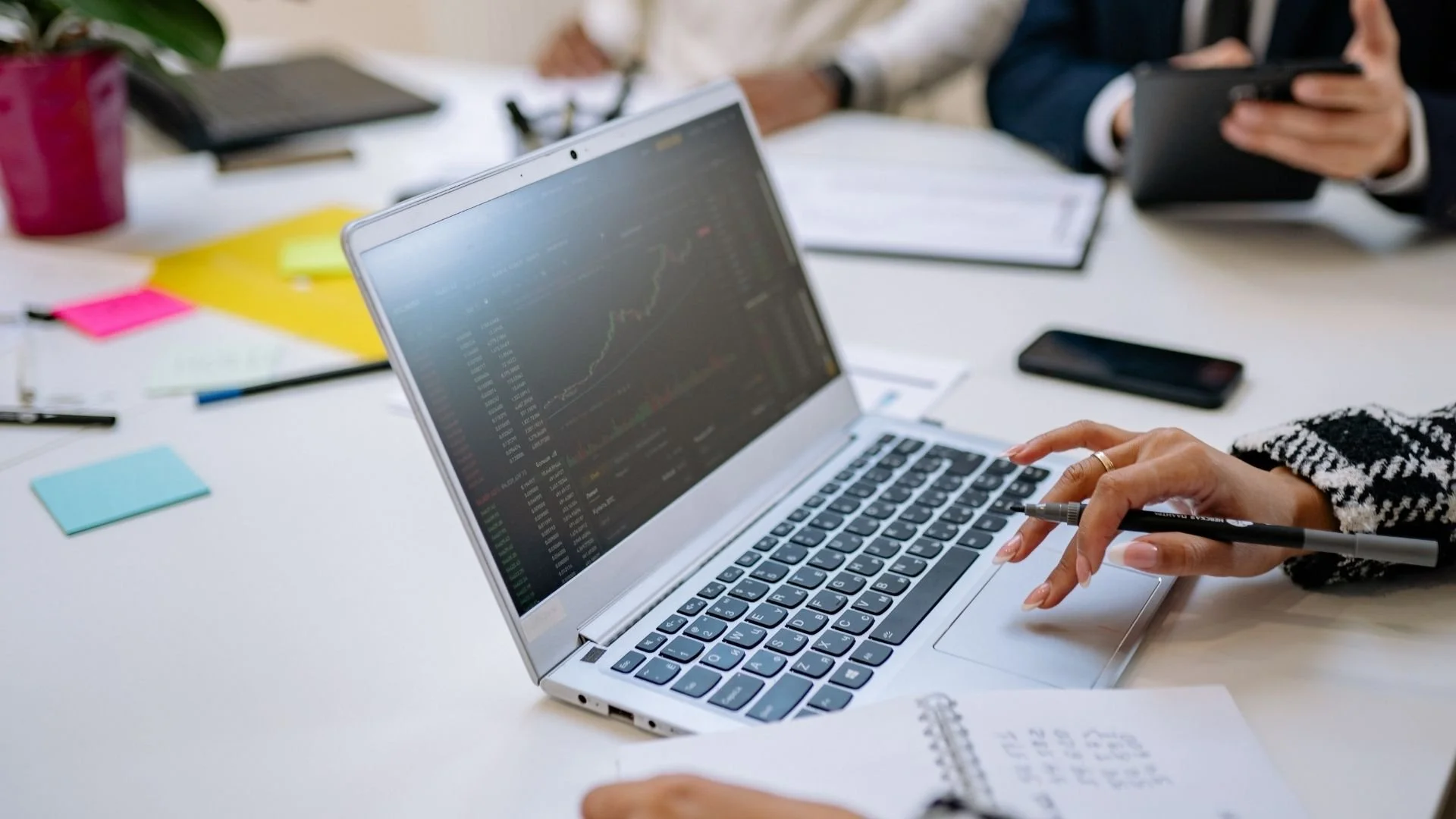Person using a laptop displaying financial charts during a team meeting, with colleagues, notes and a phone on the table.
