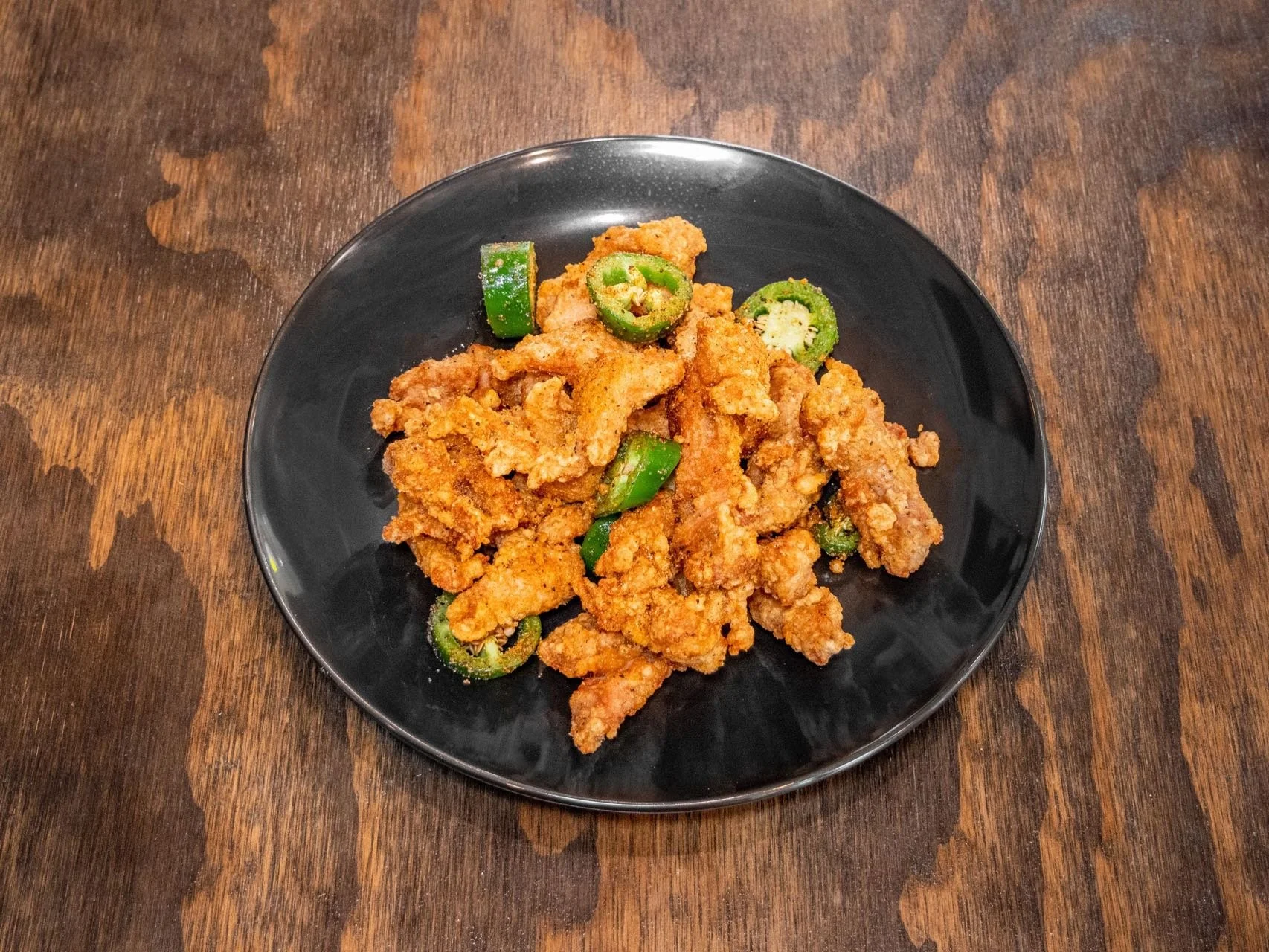 Fried chicken pieces with sliced jalapenos on a black plate resting on a wooden table.