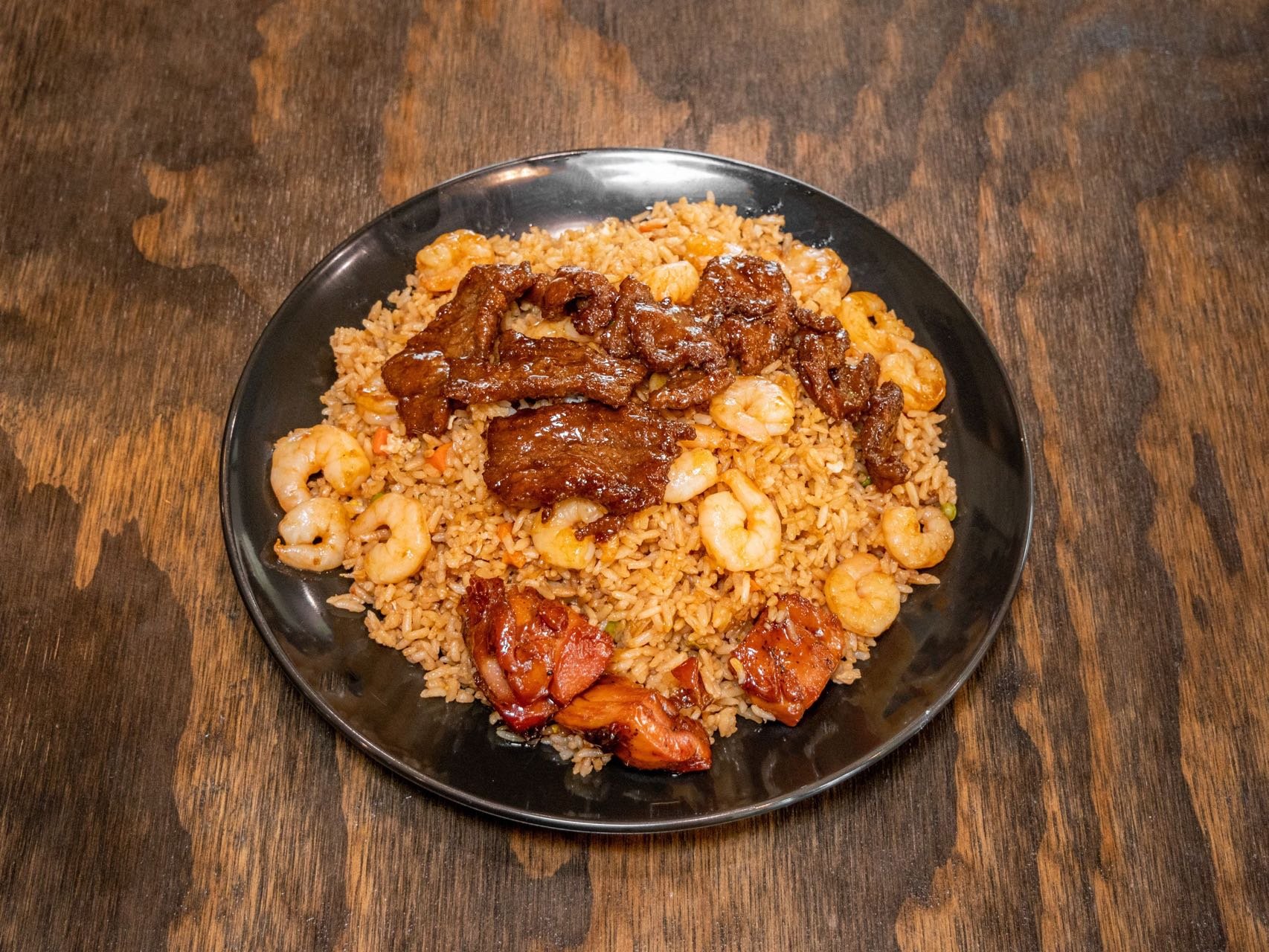 A plate of fried rice topped with beef, shrimp, and chunks of chicken. The plate is on a wooden table.