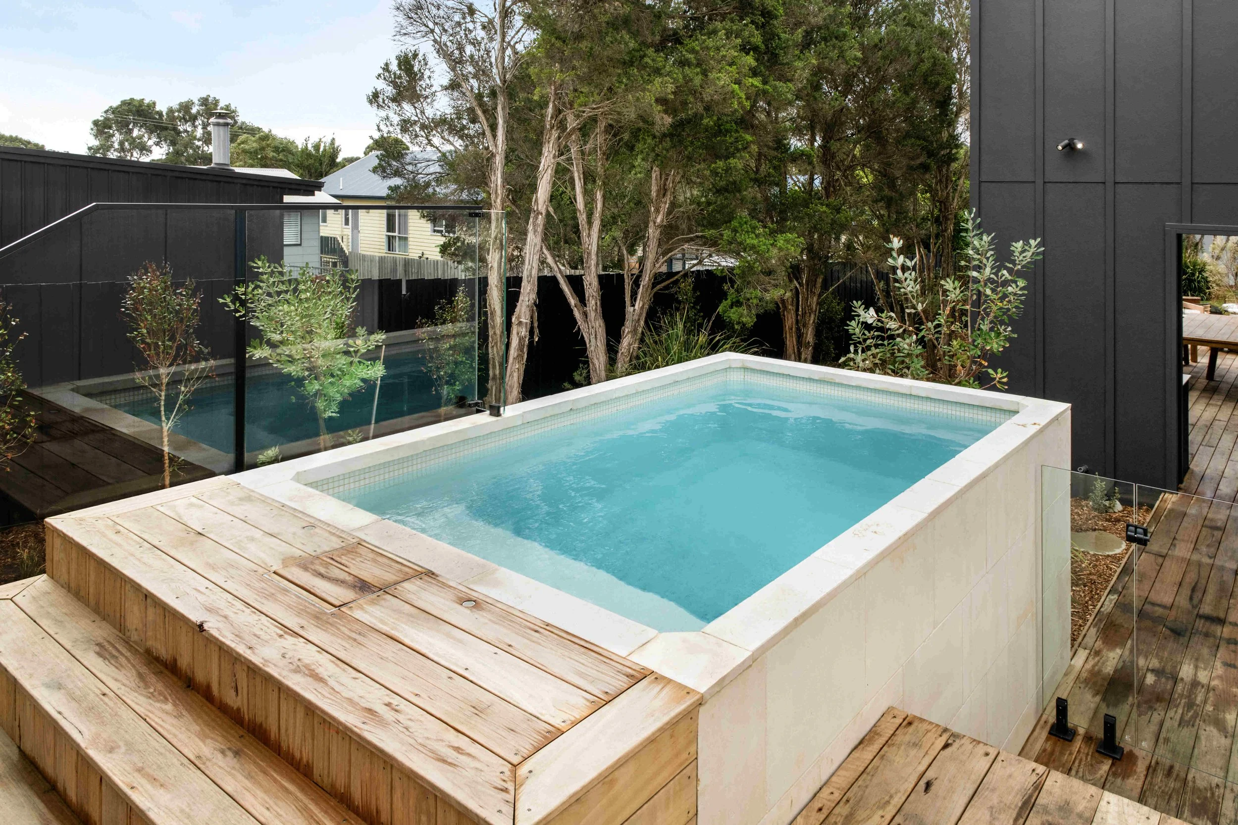 A modern outdoor hot tub with a clear glass fence, surrounded by wooden decking and greenery, with trees and neighboring houses visible in the background.