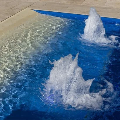 Fountains in a swimming pool with blue water and beige deck edges.