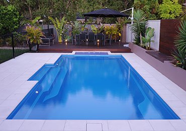 A backyard swimming pool with a deck, surrounded by lush greenery and outdoor furniture.