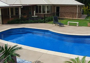 Backyard with a kidney-shaped swimming pool surrounded by concrete patio, green plants, a lounge chair, and a brick house with a porch in the background.