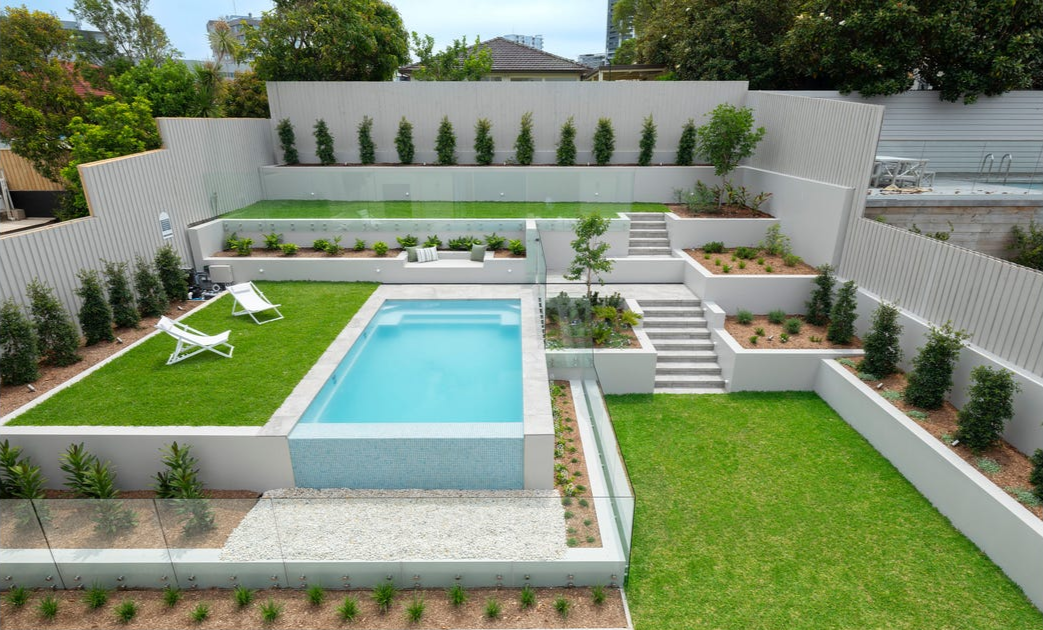 Modern backyard with a swimming pool, green lawns, lounge chairs, and tiered garden beds with trees and shrubs, surrounded by a white fence.