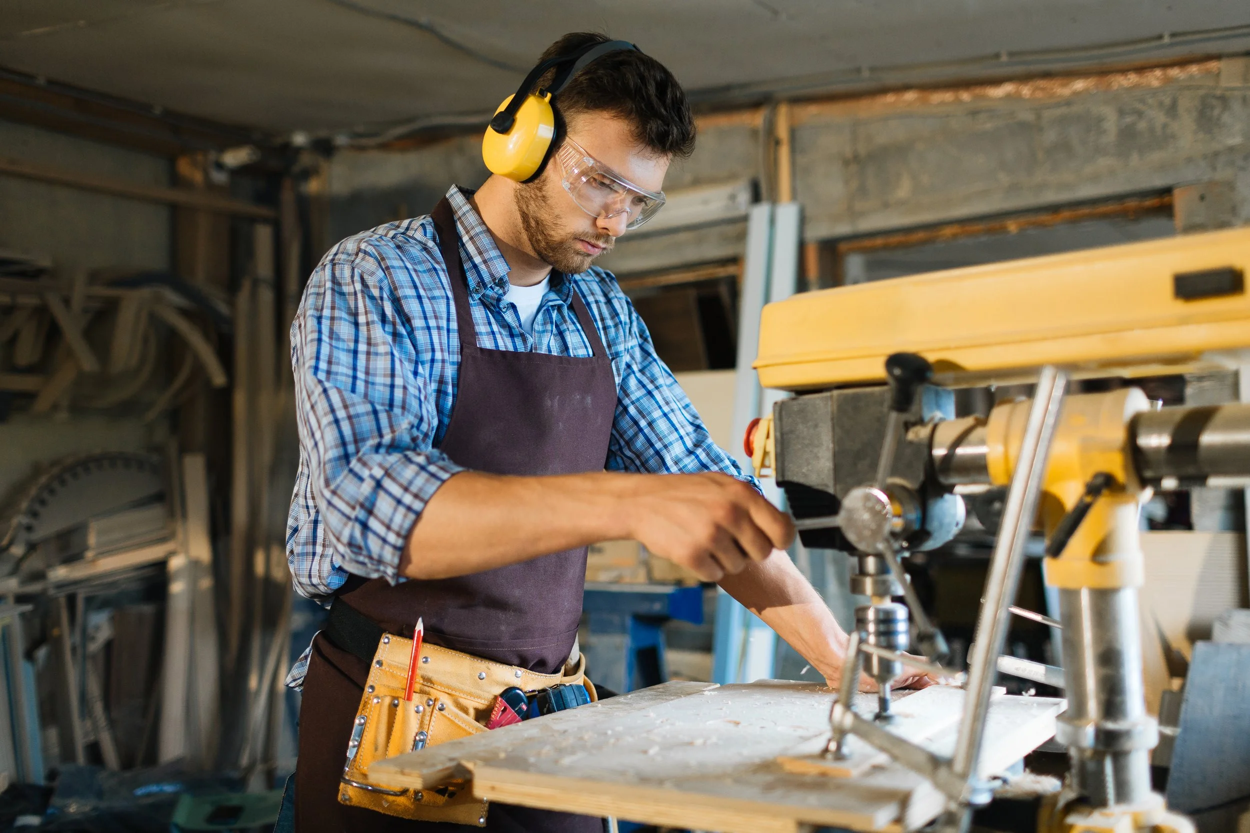 Man working in a woodworking shop, operating a drill press, wearing ear protection and safety goggles, with woodworking tools attached to his apron.